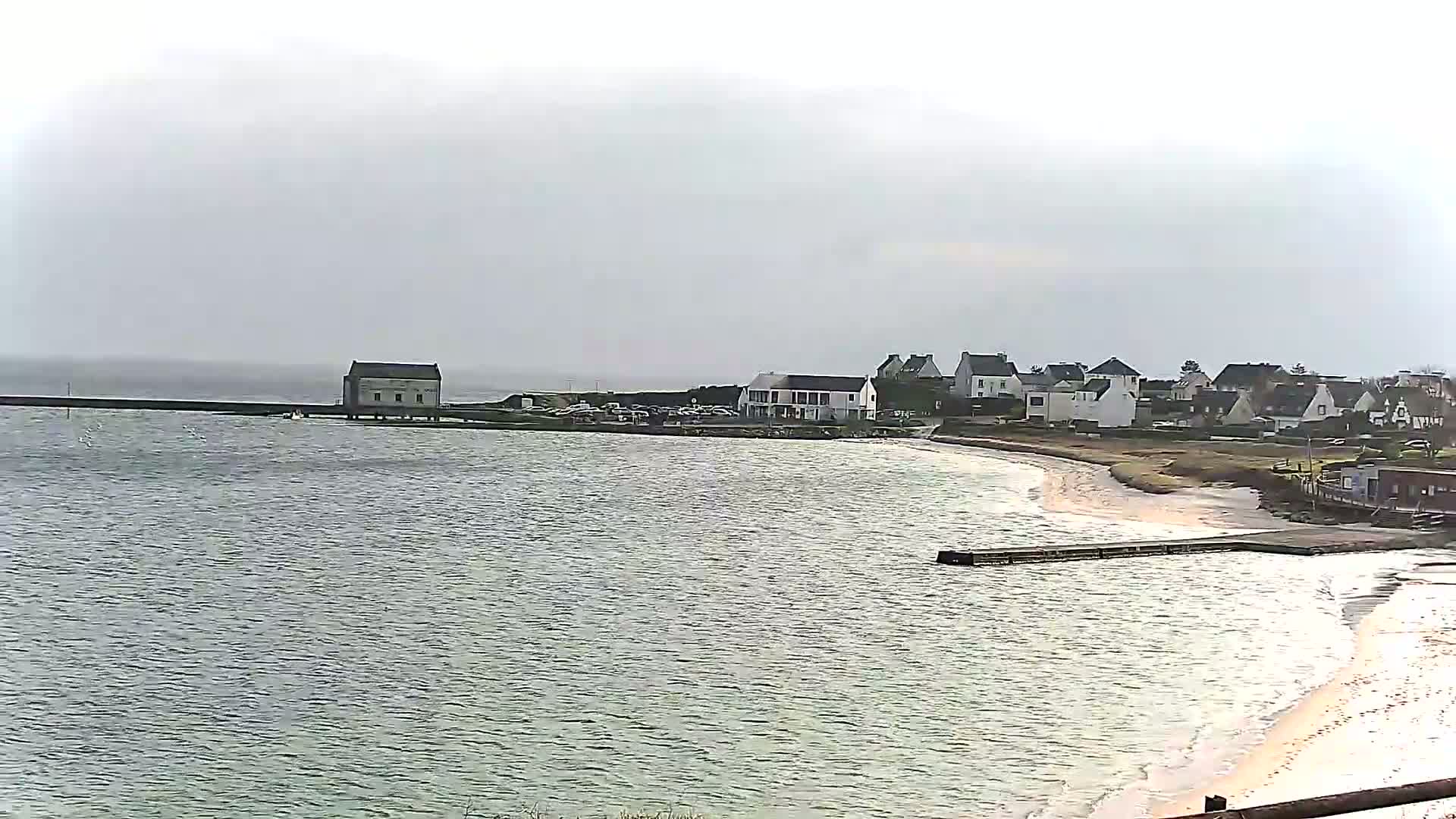 A calm body of water laps a sandy shore next to a small village under an overcast sky.