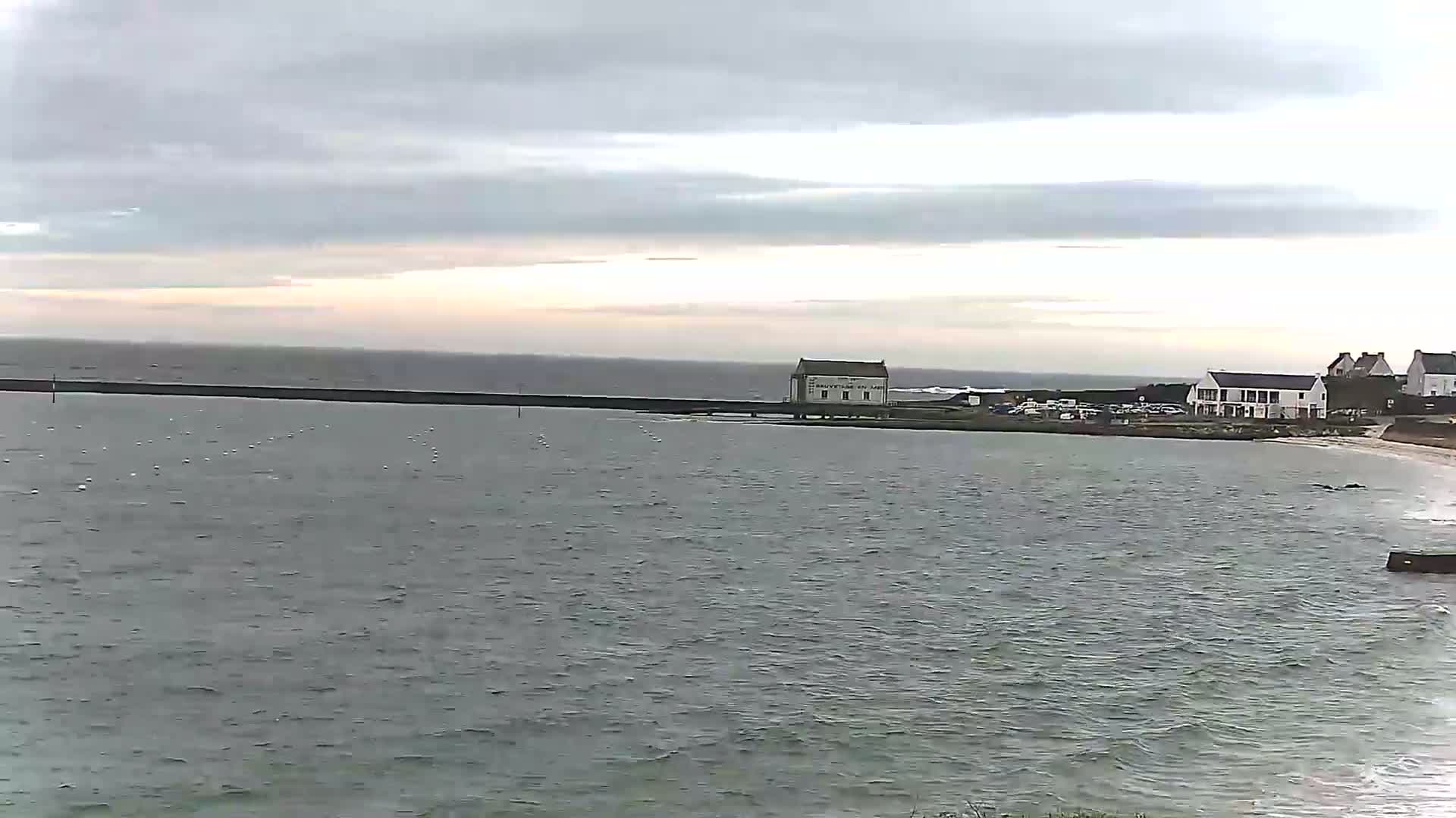 A calm, gray day shows a body of water with a long seawall and a few buildings on the shore, including a small white building near the water's edge.