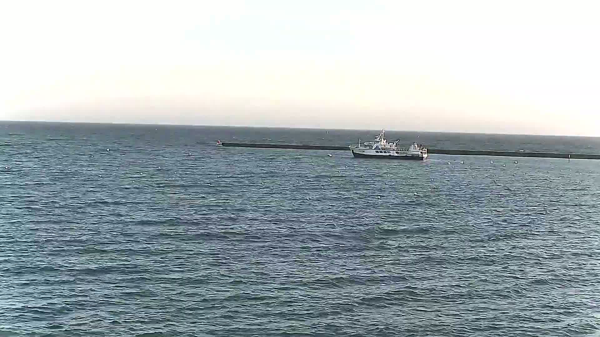 A passenger boat is anchored near a long, dark breakwater in a calm sea under a pale sky.
