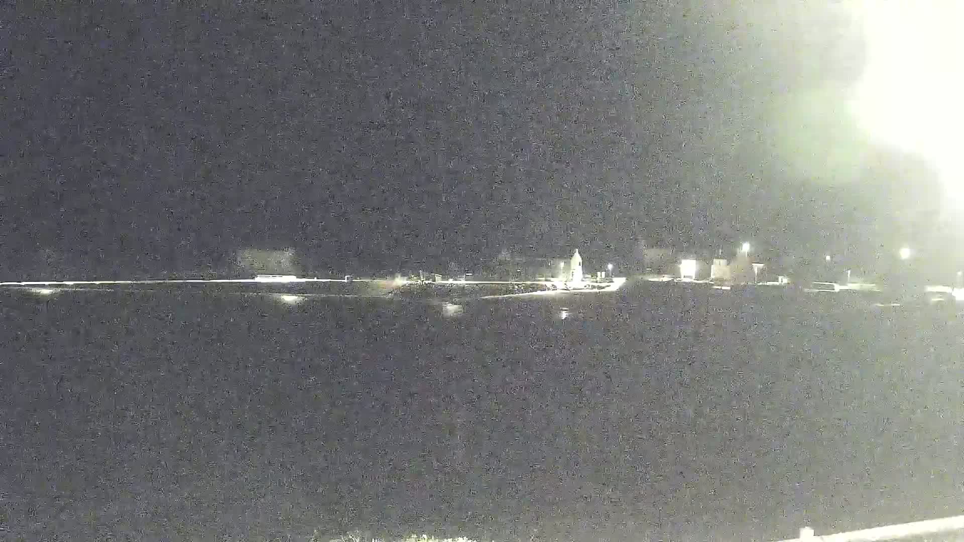 A nighttime, long shot of a dark body of water reflecting the lights of a distant, sparsely lit shoreline.