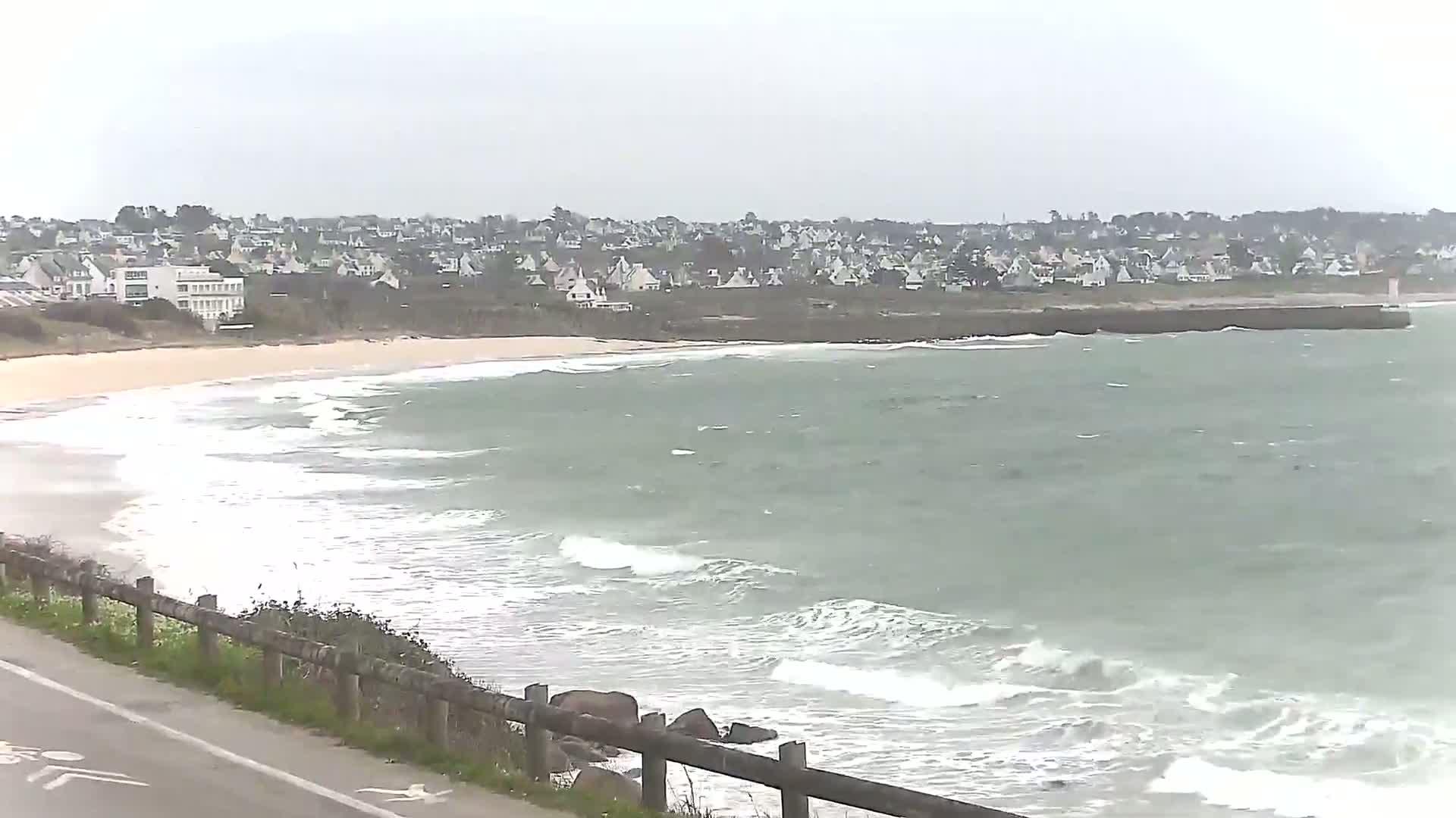 A sandy beach with small waves and a town of houses on a hill overlooking it on an overcast day.