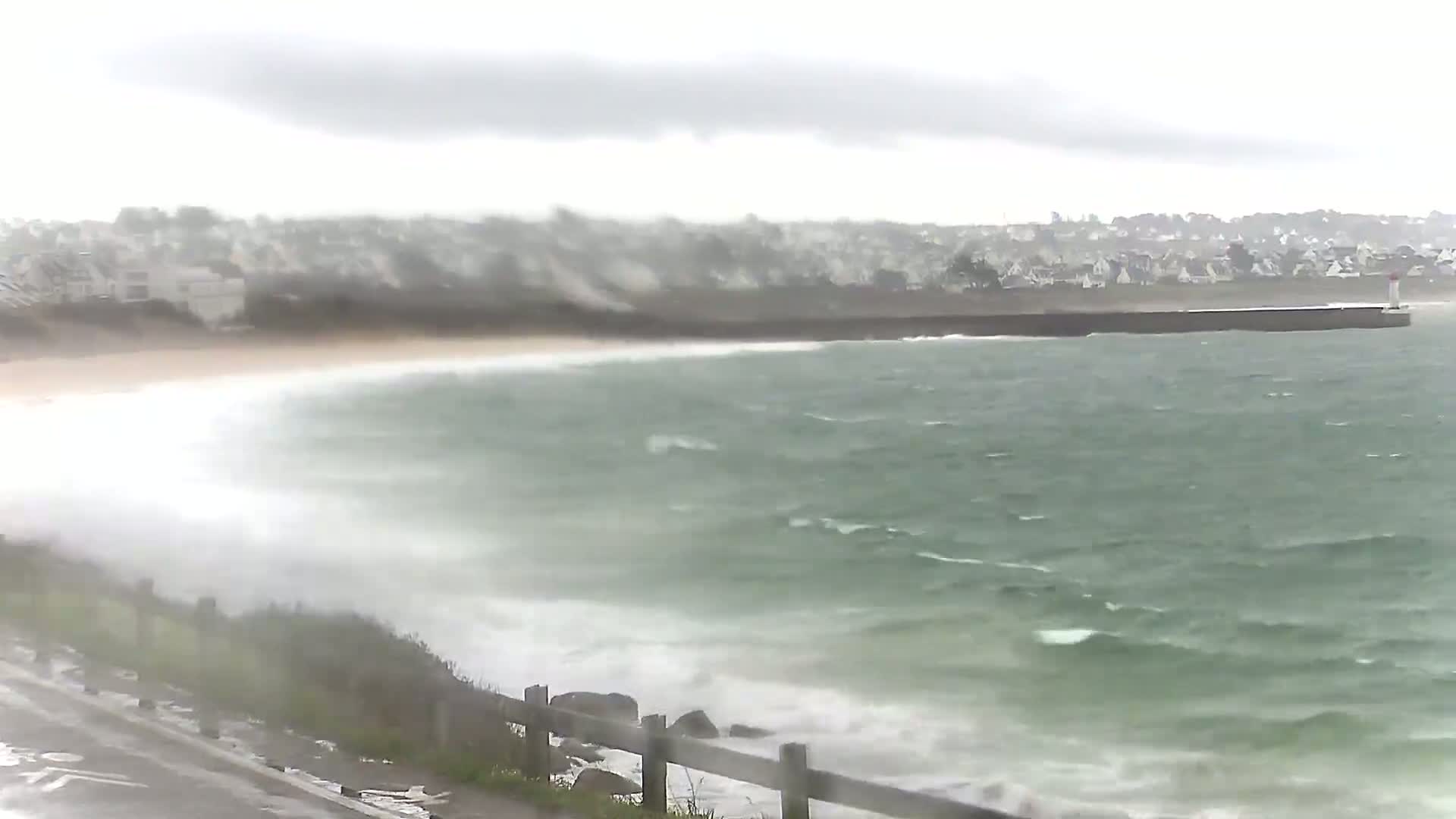 A coastal scene shows a stormy ocean with choppy waves crashing on a sandy beach near a residential area under an overcast sky.