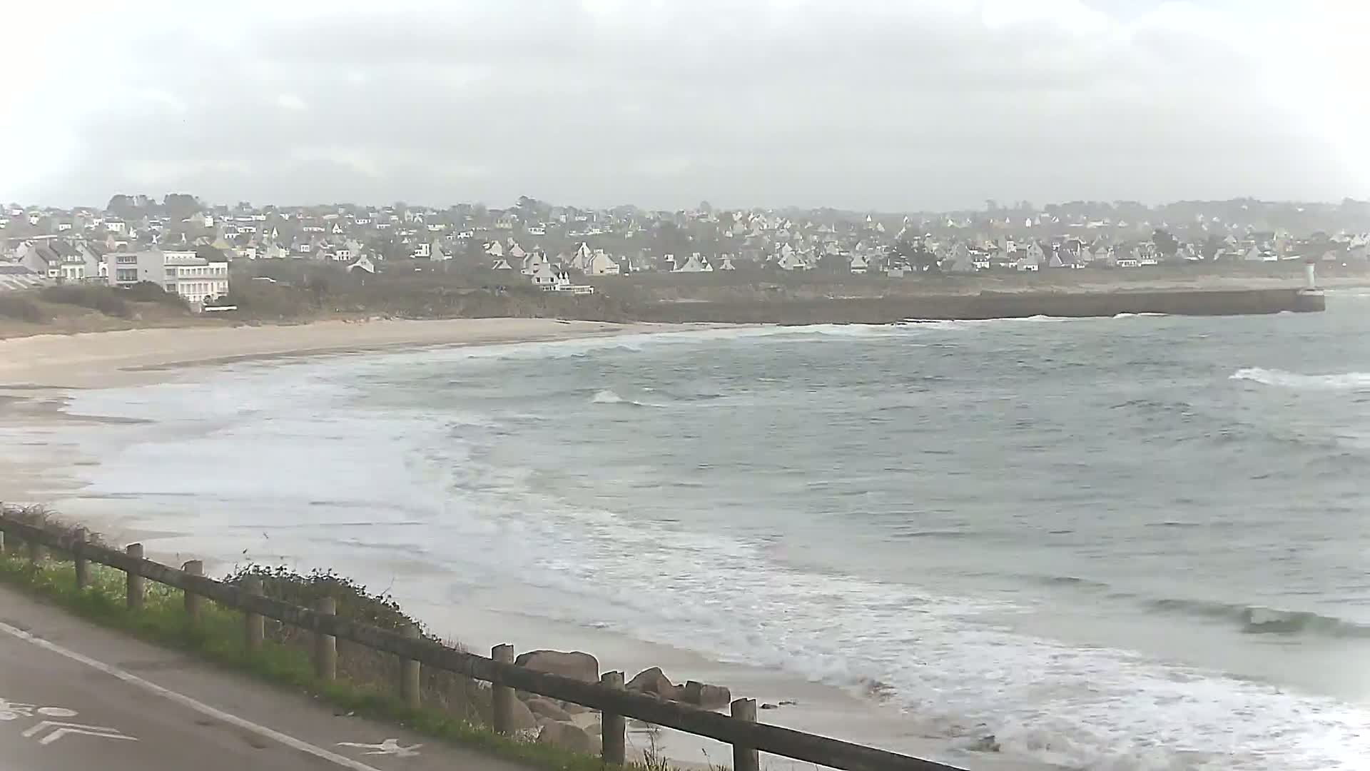 A sandy beach with gentle waves laps the shore under an overcast sky, with a town visible in the distance.