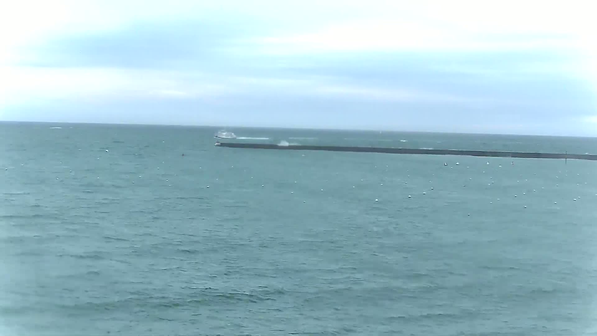 A boat passes a long, dark breakwater in a choppy sea under a cloudy sky.