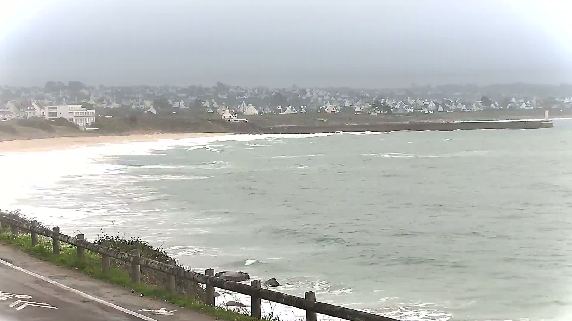 A coastal road overlooks a gray, overcast ocean with small waves breaking on a sandy beach and a distant town.