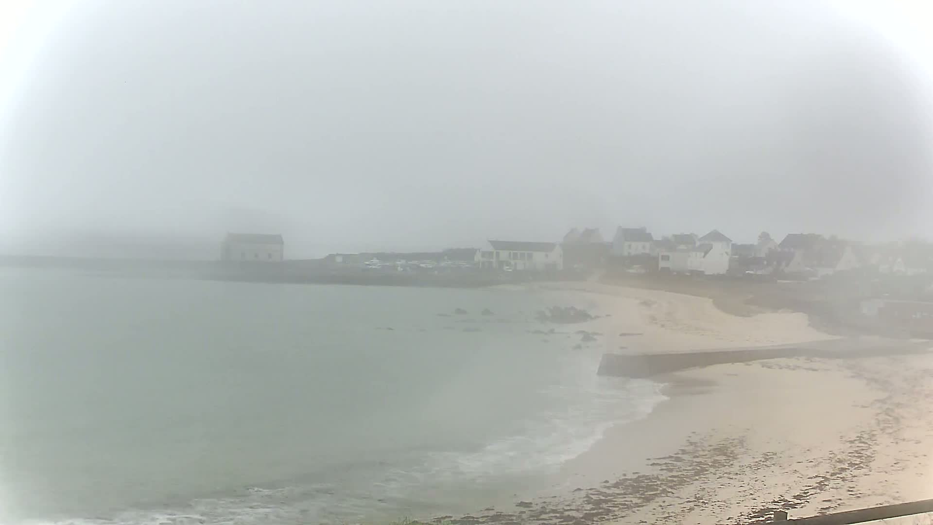 A foggy coastal scene shows a small village near a sandy beach and calm water.