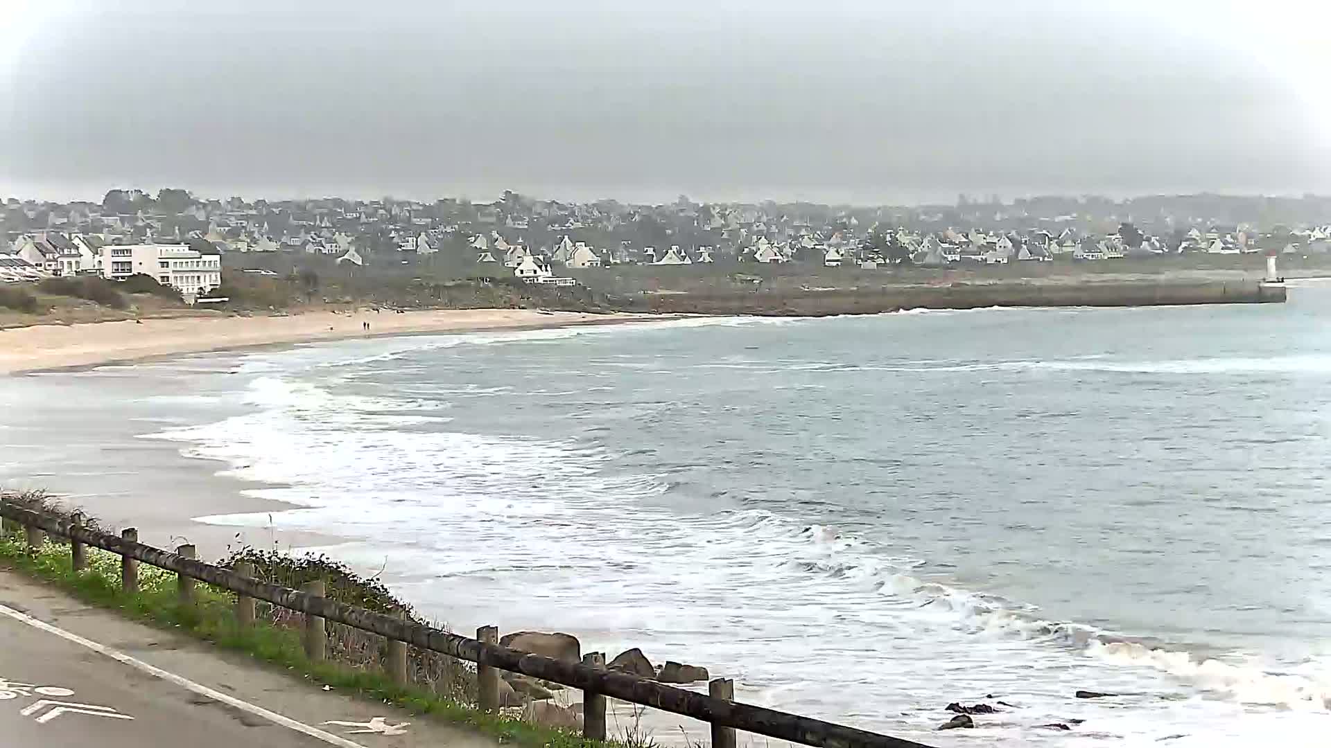 A sandy beach with gentle waves under an overcast sky is seen from a roadside vantage point, overlooking a town in the distance.