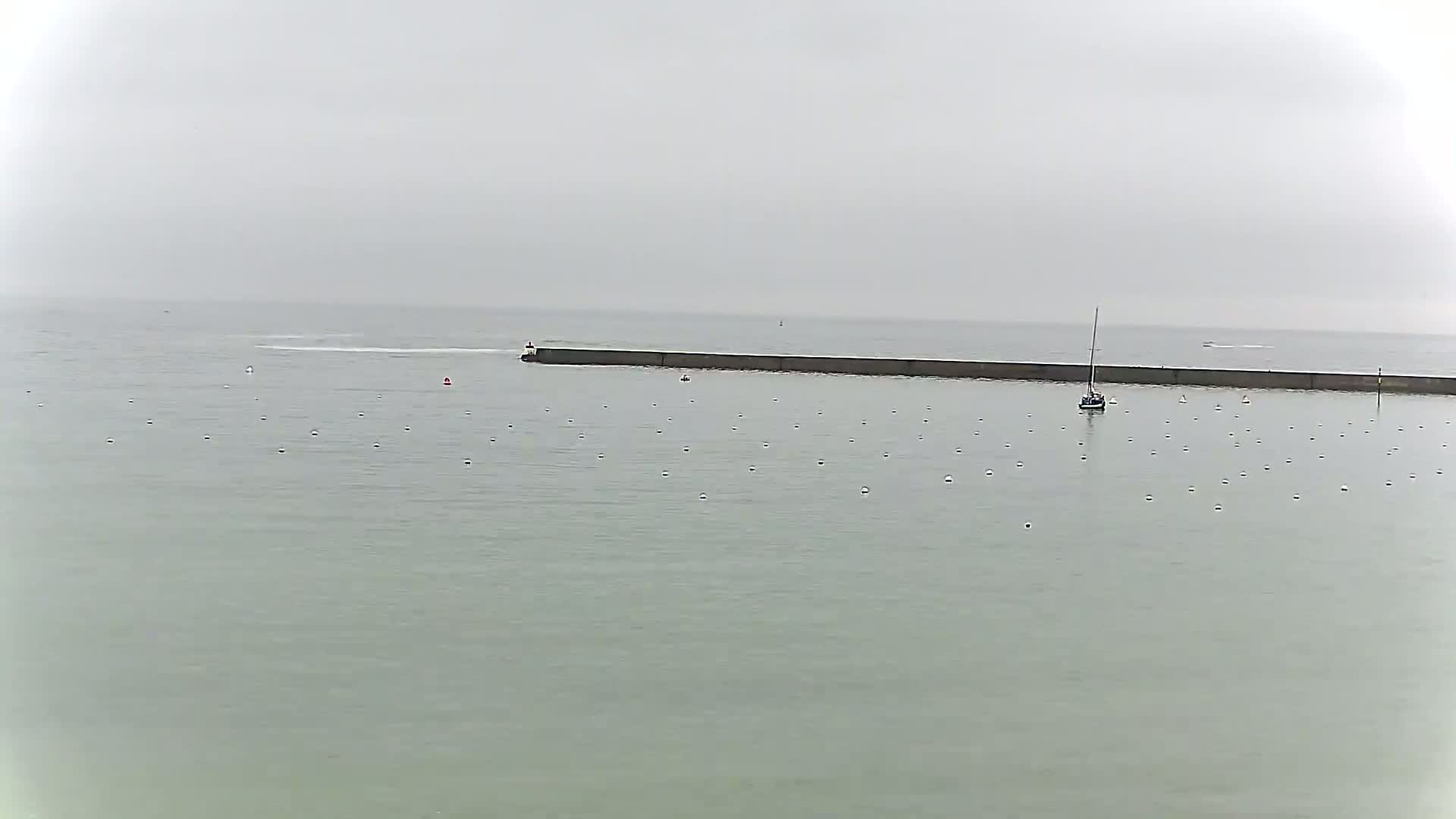 A long, low seawall stretches across a calm, gray sea under an overcast sky, with a sailboat and numerous white buoys visible in the water.