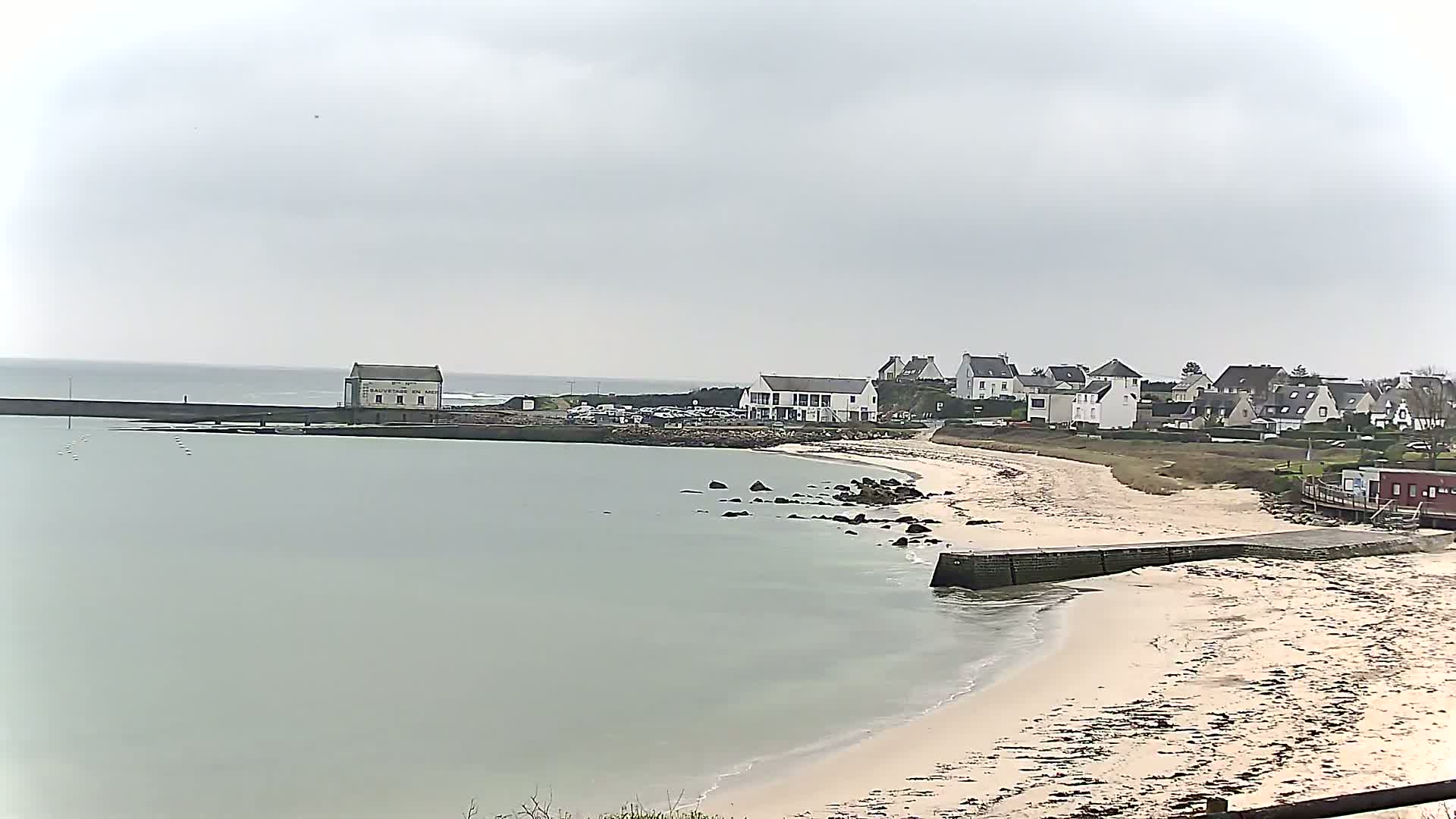 A sandy beach curves around calm, light-green water toward a small town under an overcast sky.