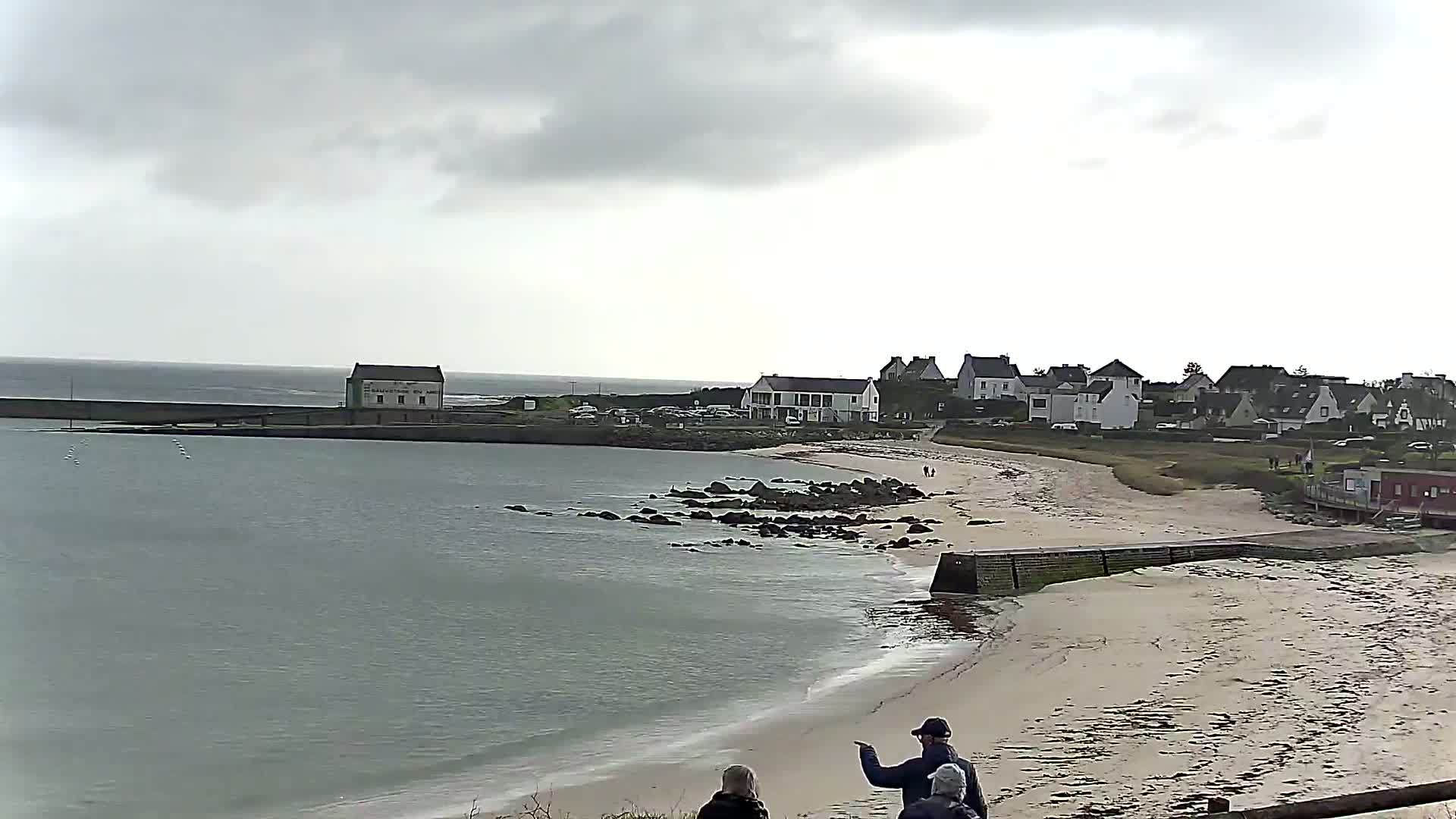 A calm, overcast day reveals a sandy beach, a small town, and a calm bay with a few rocks near the shore, all viewed from a slightly elevated vantage point.