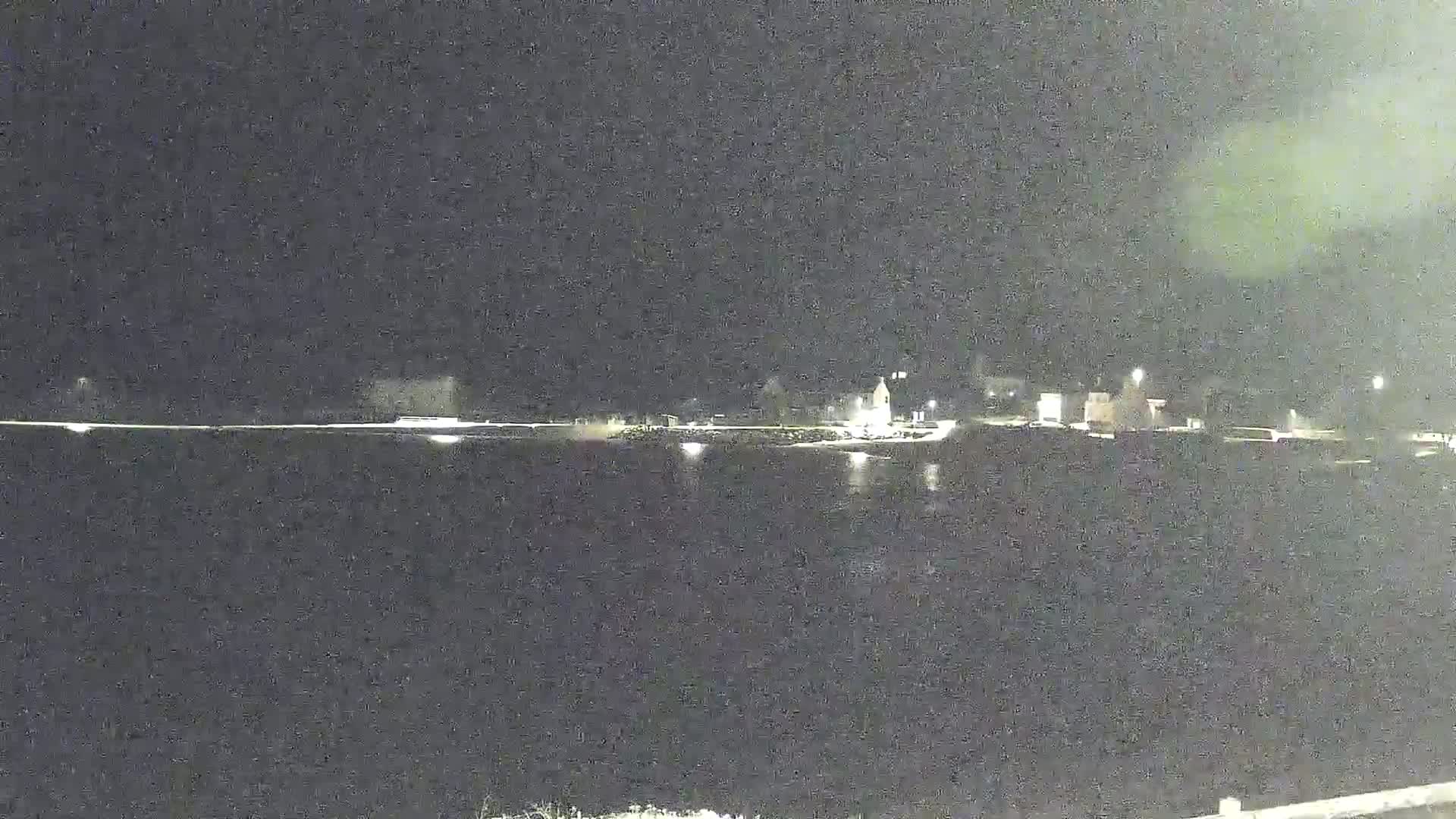 A nighttime view shows a dark body of water reflecting the lights of a distant shoreline with buildings.