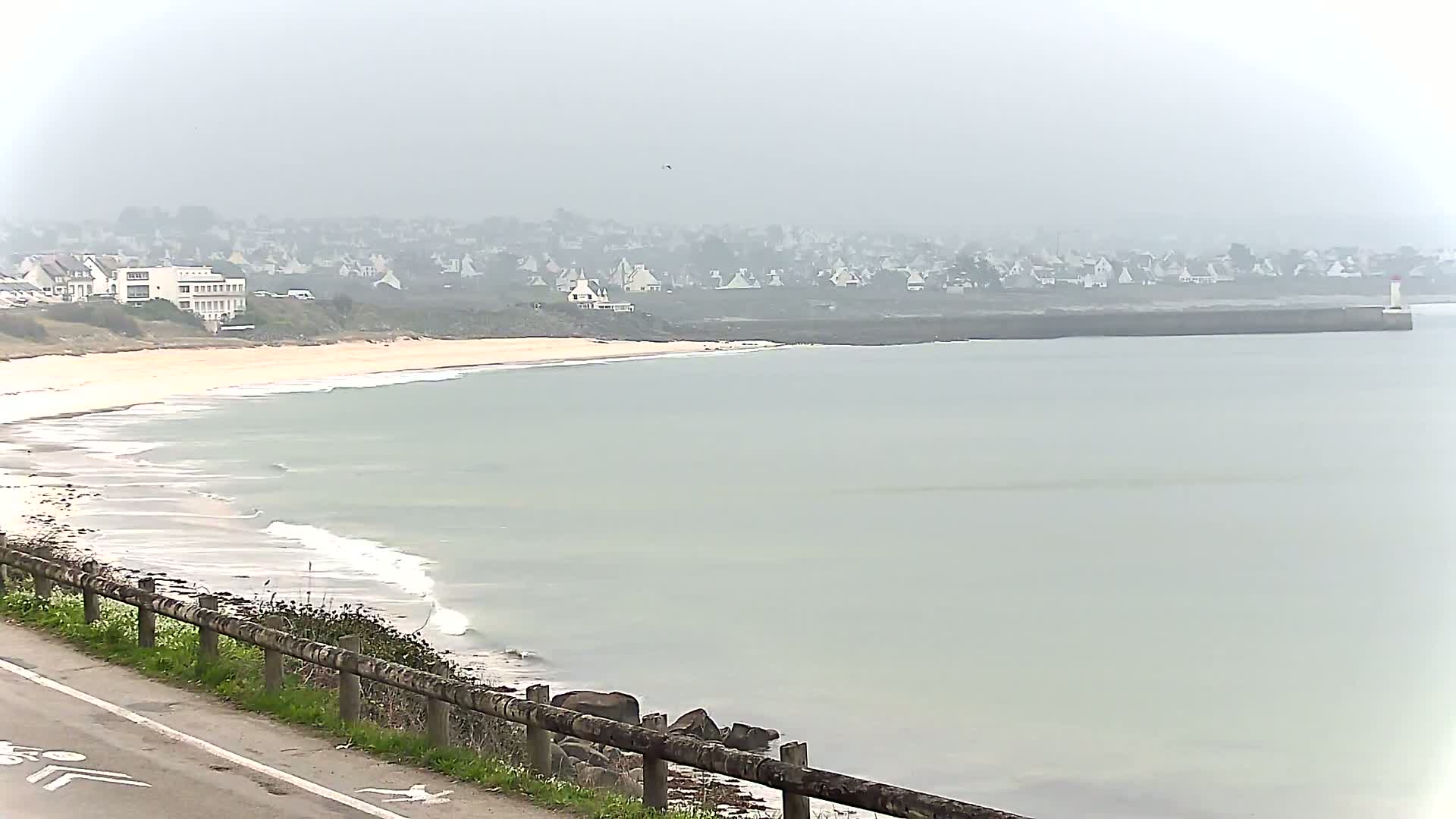 A sandy beach with gentle waves laps the shore under a foggy sky, with a distant town and breakwater visible.