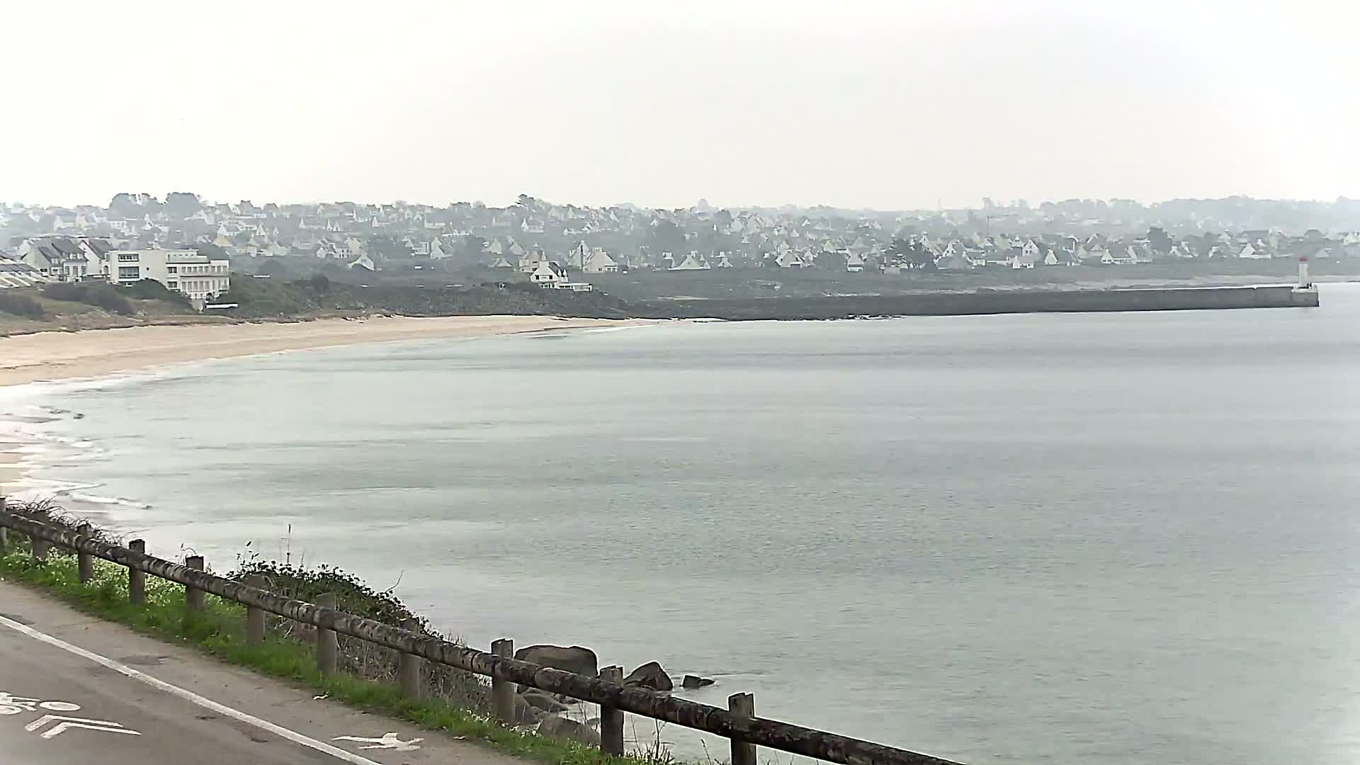 A sandy beach curves along a calm, gray sea under an overcast sky, with a town of low buildings visible in the distance and a small lighthouse on a breakwater.