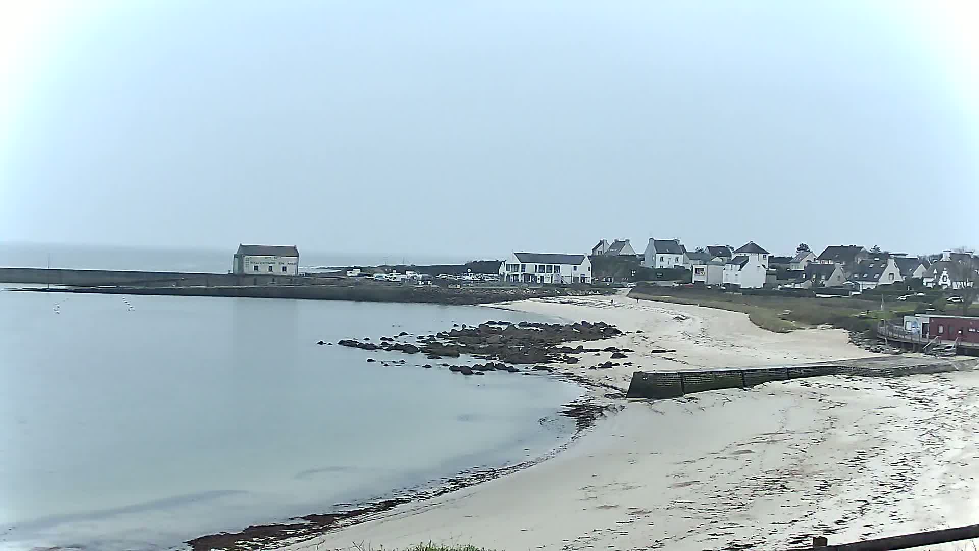 A sandy beach and calm ocean water meet a small village of white buildings under an overcast sky.