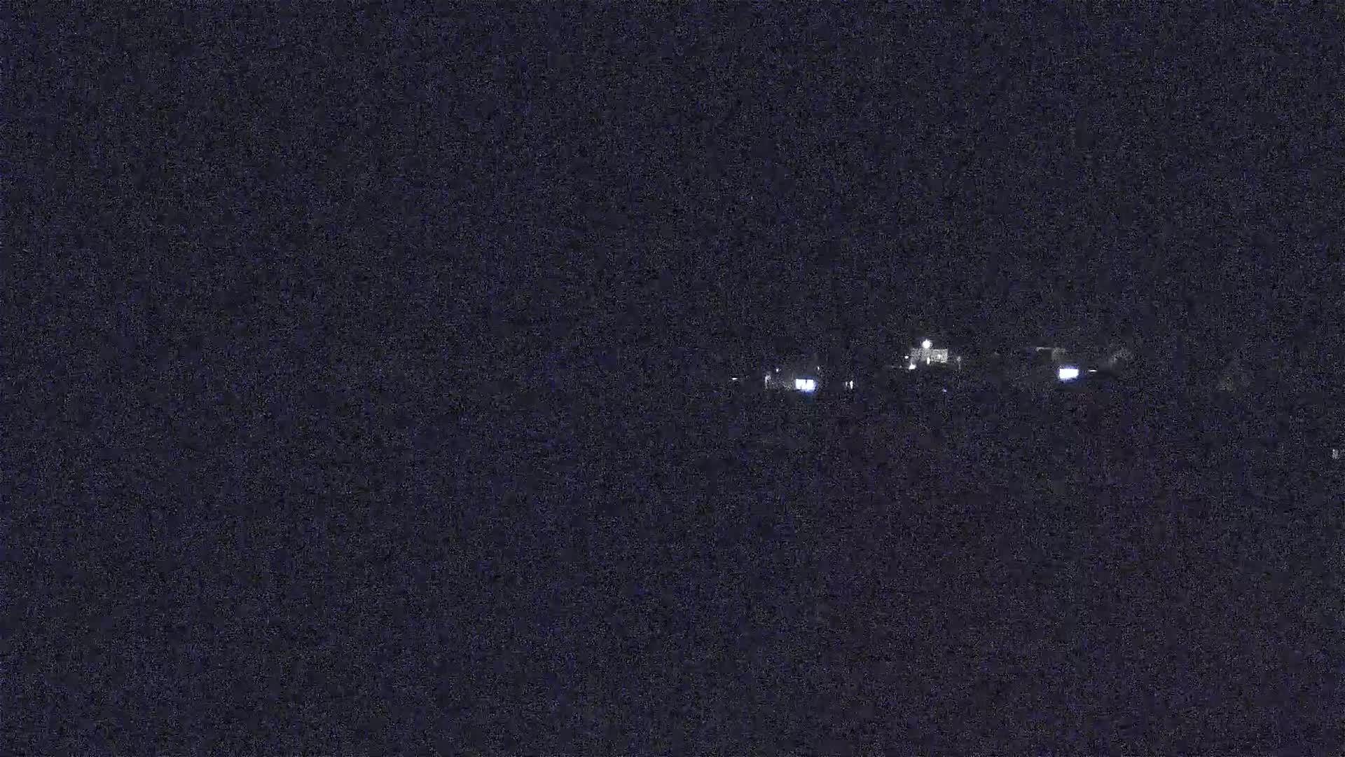 A nighttime long shot shows a few faintly lit buildings on a dark horizon under a clear, dark sky.