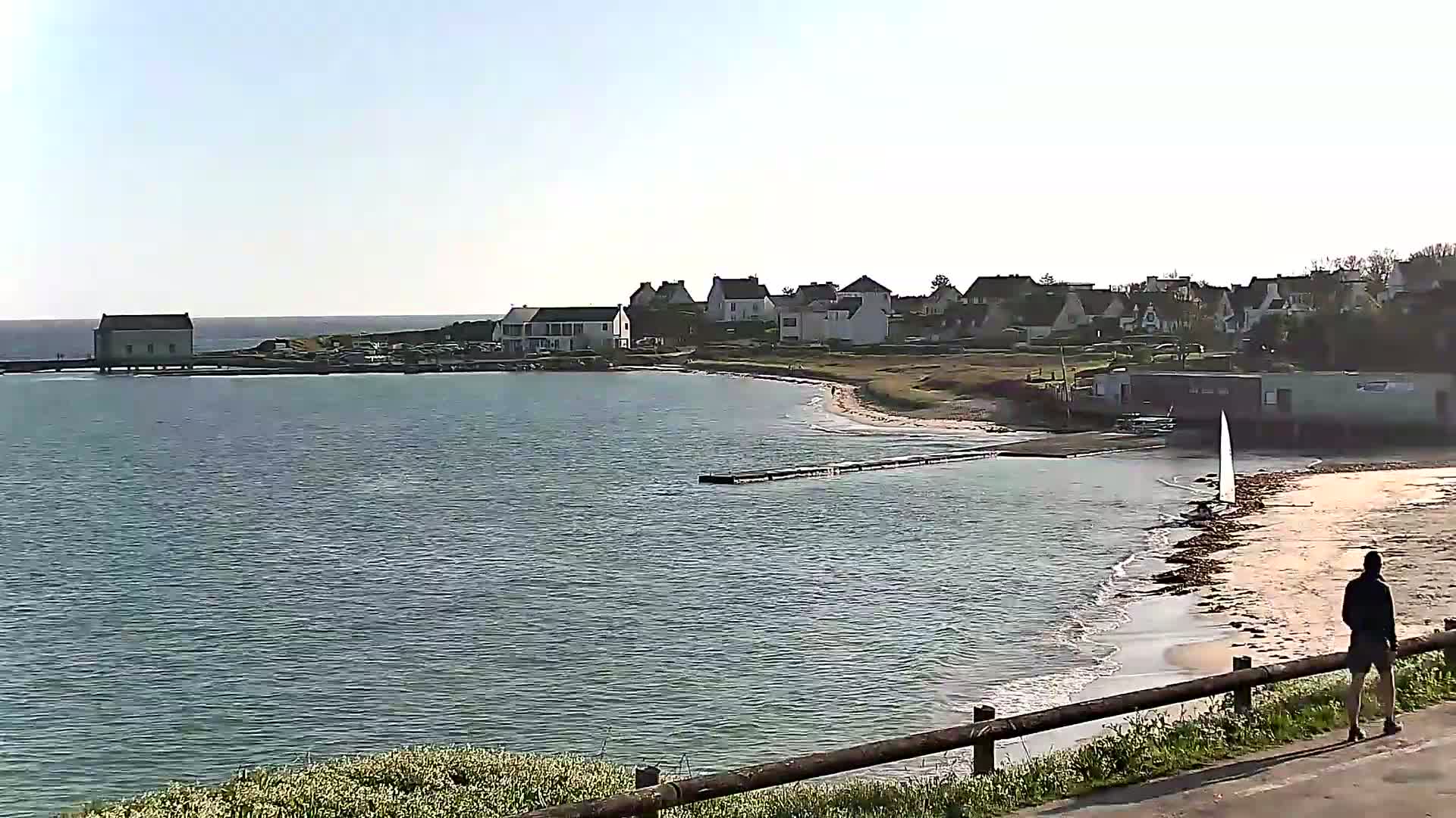 A calm body of water laps a sandy shore near a small village under a clear, bright sky; a lone person stands on a path overlooking the scene.