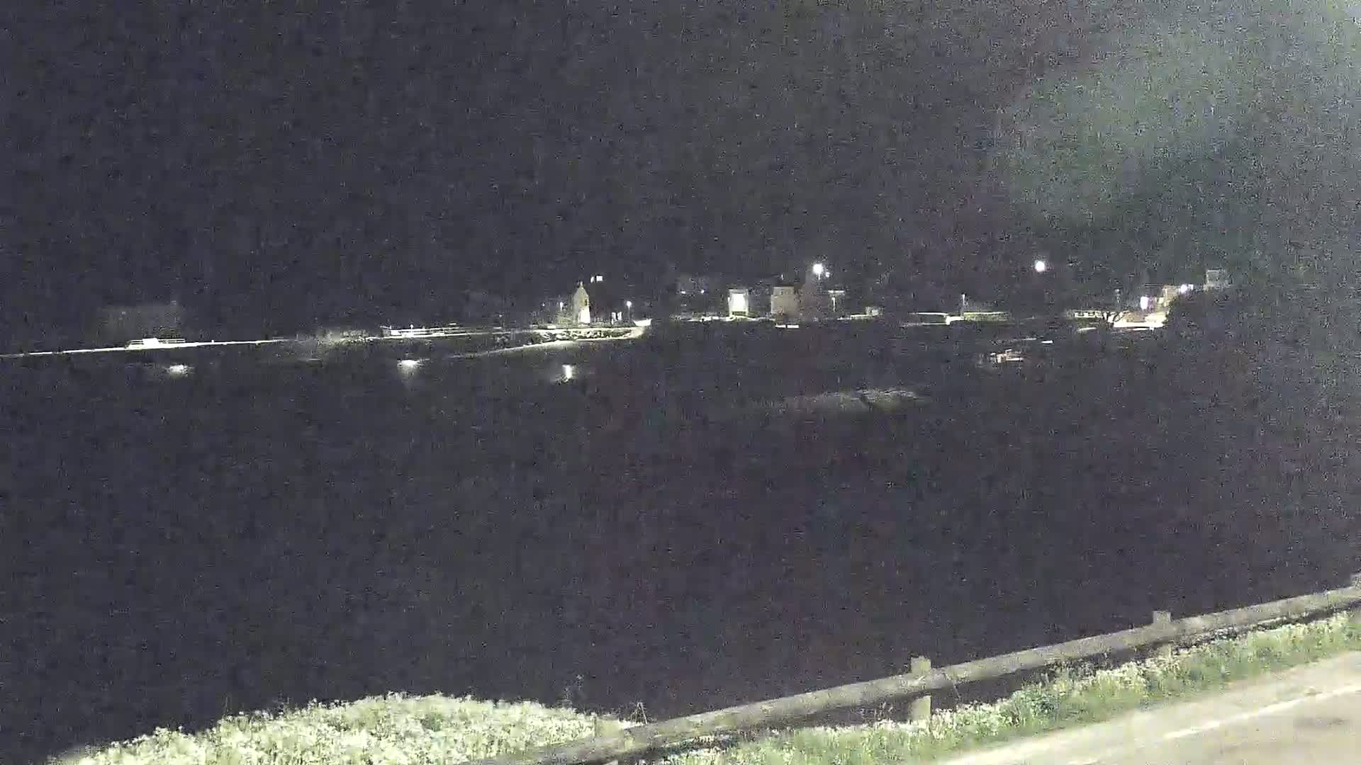 A nighttime view of a calm body of water with dimly lit buildings and vegetation visible in the background under a dark sky.