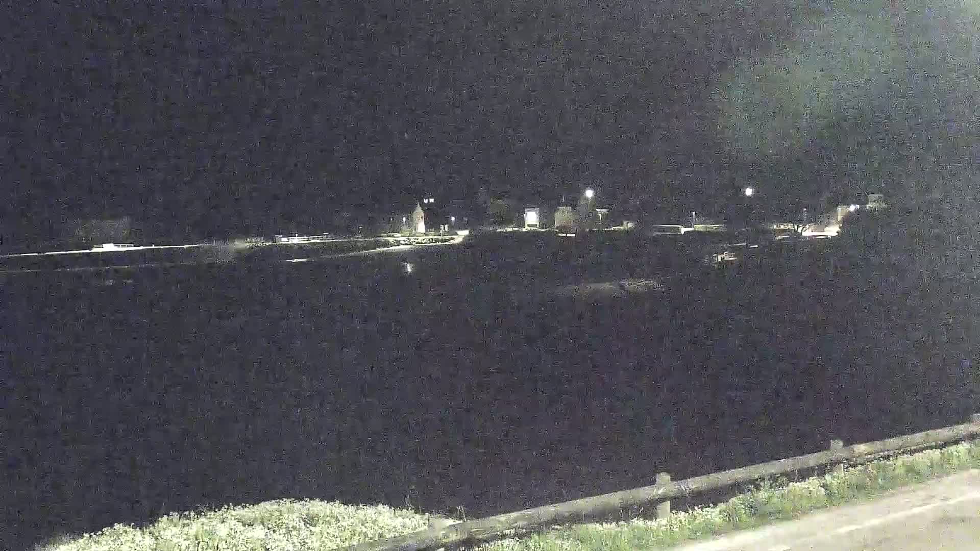 A nighttime view of a dark body of water, sparsely lit buildings in the distance, and a grassy area with a fence in the foreground.