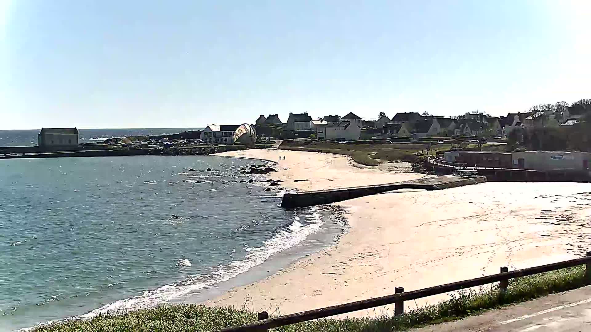 A sandy beach curves into a calm, light-blue sea next to a small village under a clear, sunny sky.