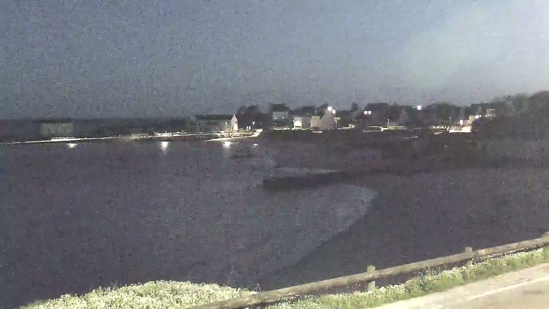 A dark, calm body of water stretches out before a dimly lit town under a dusky sky.