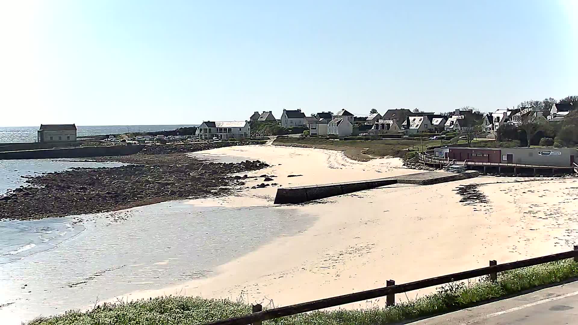 A sandy beach curves around a rocky shoreline, leading to a small village of houses under a clear, sunny sky.