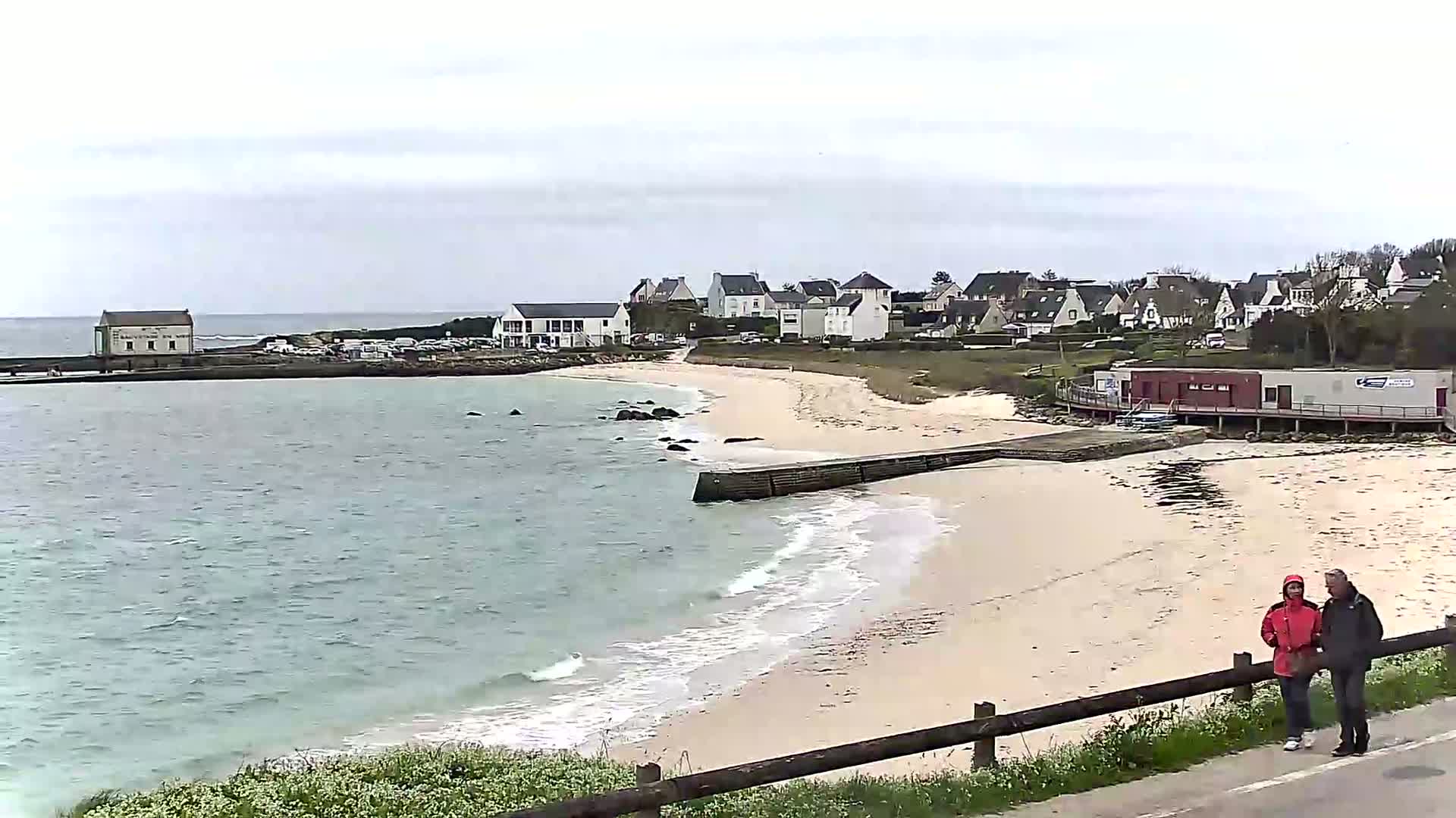 A sandy beach with gentle waves laps the shore near a small village under an overcast sky, with a couple walking along the road above.