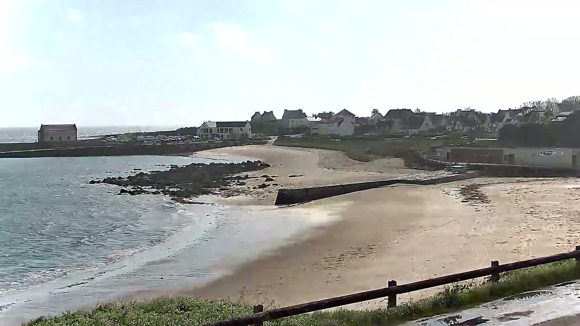 A sandy beach curves around a small coastal town under a mostly sunny sky, with calm water lapping at the shore.