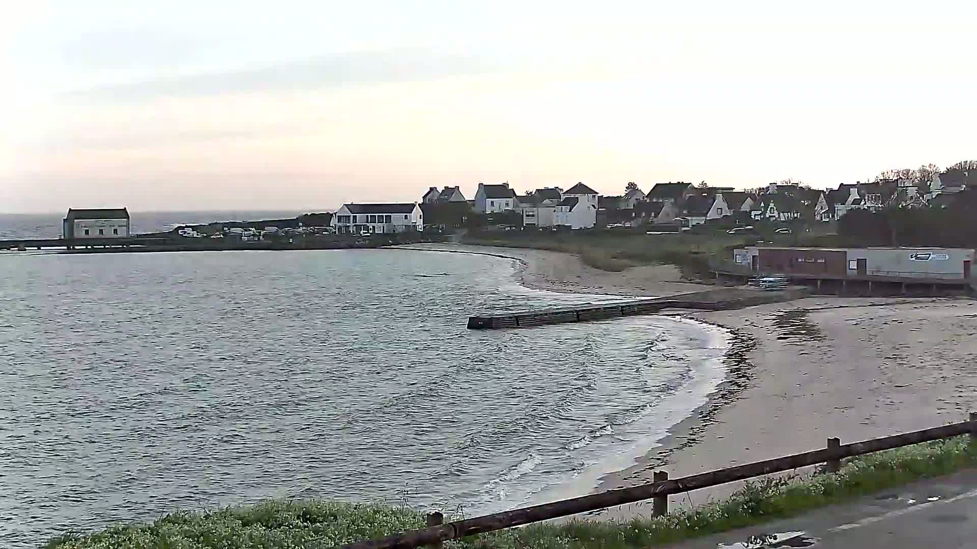 A calm, gray day shows a small coastal town nestled beside a sandy beach and a body of water, with gentle waves lapping the shore.