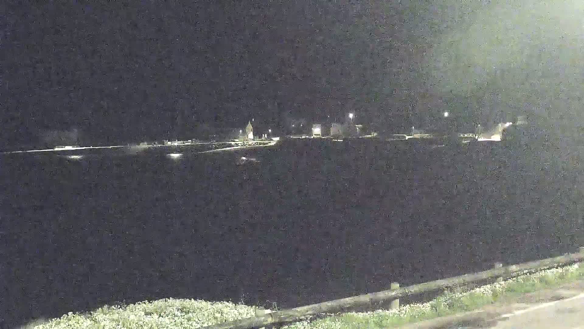 At night, a dimly lit landscape is visible from a slightly elevated vantage point, showing a flat expanse of land with a few structures in the distance.