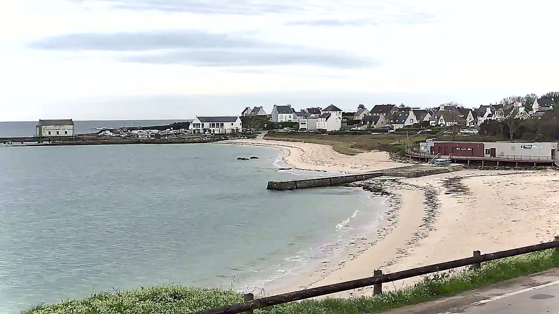A sandy beach curves around a calm bay, with a small village of white houses situated on a nearby hill under an overcast sky.