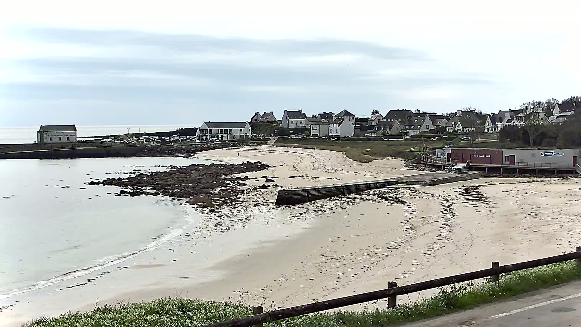 A sandy beach with calm water, rocky shoreline, and a small village of houses in the background under an overcast sky.