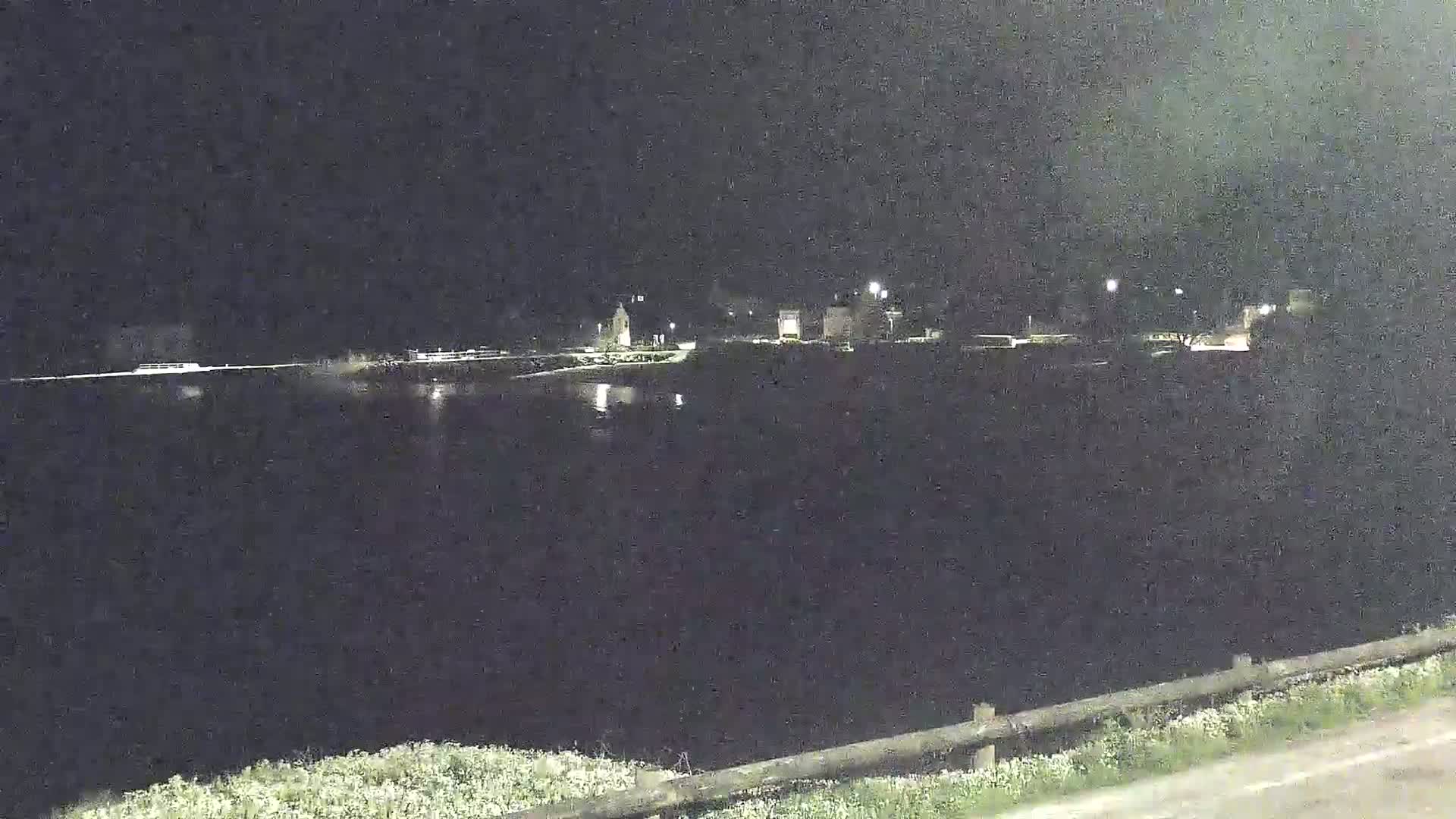 A dark, nighttime scene shows a body of water reflecting a few lights from buildings across the water, with vegetation along the bottom of the image.