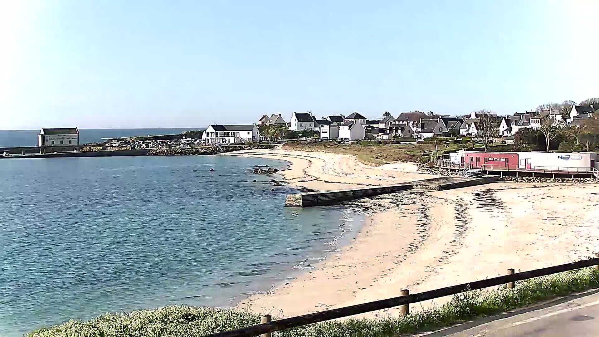 A sandy beach curves around a calm body of water, with a small village and several buildings situated on a slight incline above the shore under a clear, sunny sky.