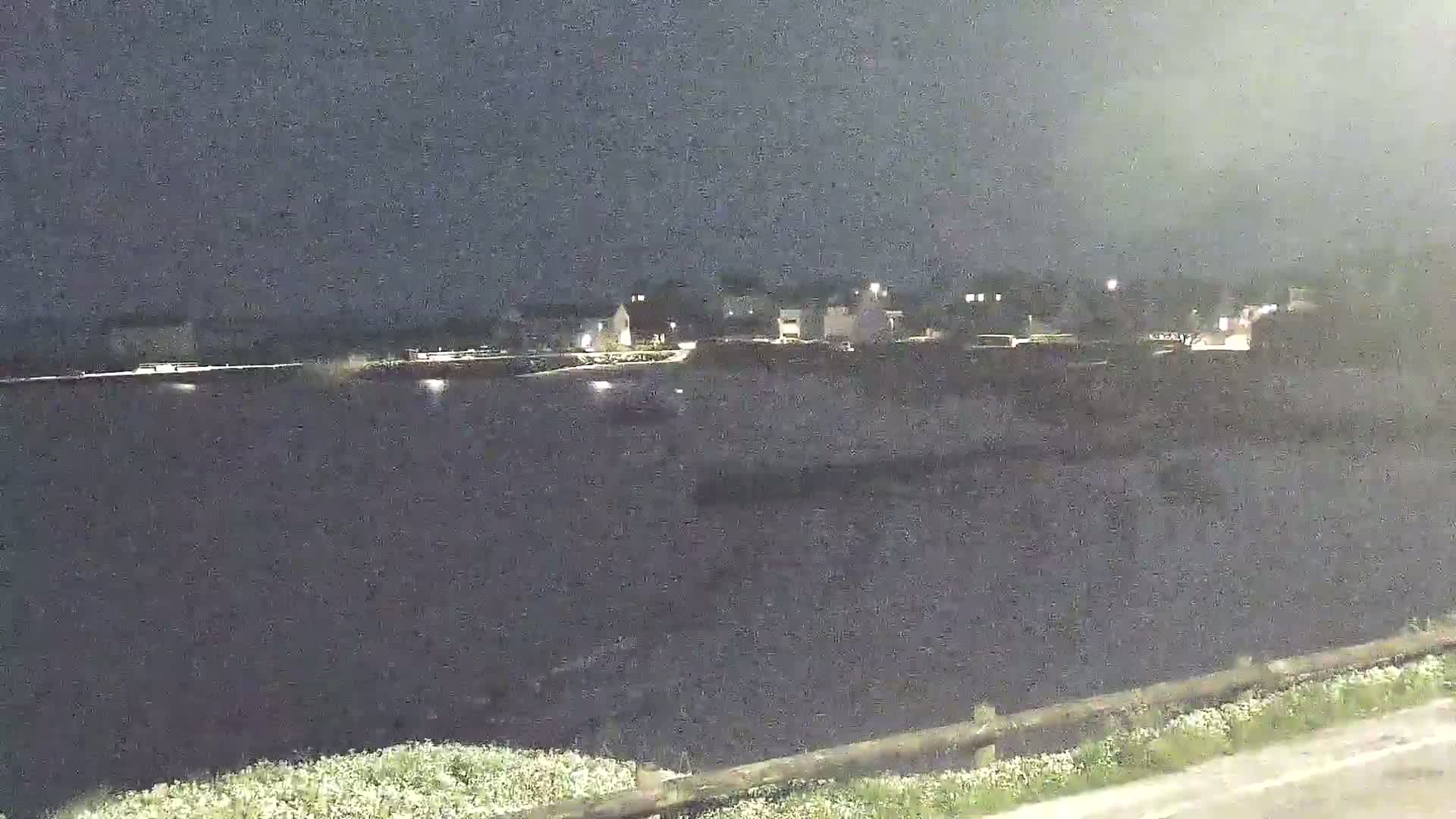A nighttime view of a dark body of water with a small town faintly lit on the opposite shore under a cloudy sky.