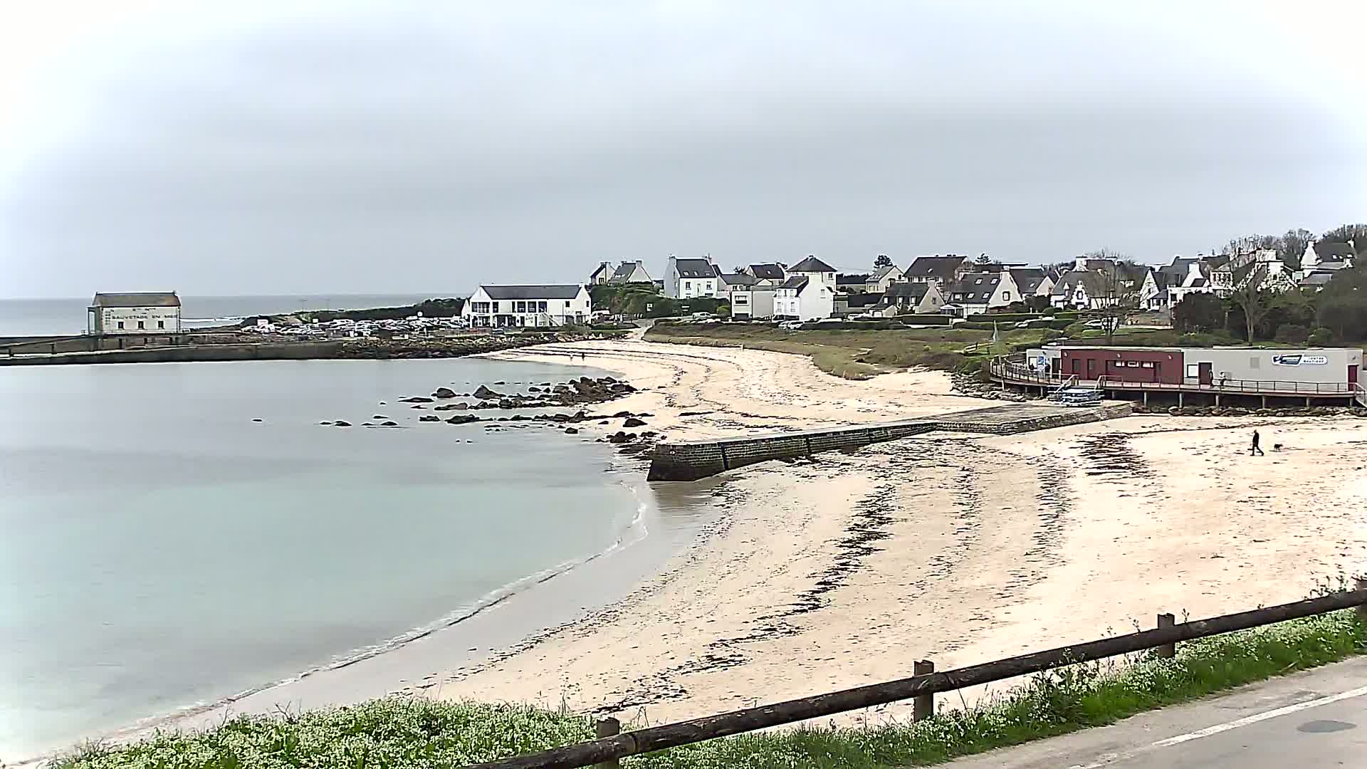 A sandy beach curves around a calm bay,  with a small village of white buildings behind it, under an overcast sky.