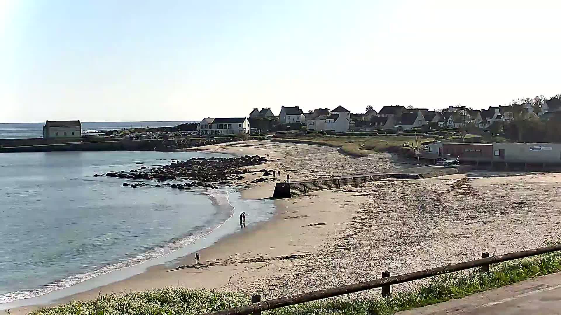 A sandy beach with calm water laps gently at the shore, overlooking a small coastal village under a clear, sunny sky.