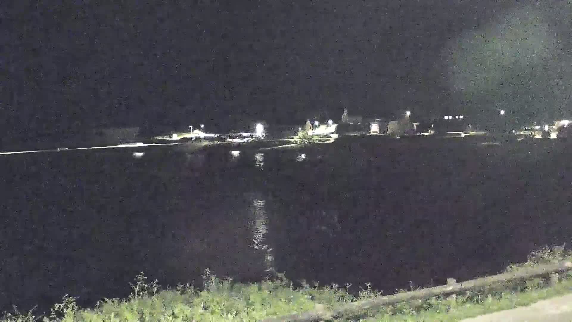A nighttime view shows a body of dark water reflecting some lights from a small town across the water, under a clear, dark sky.