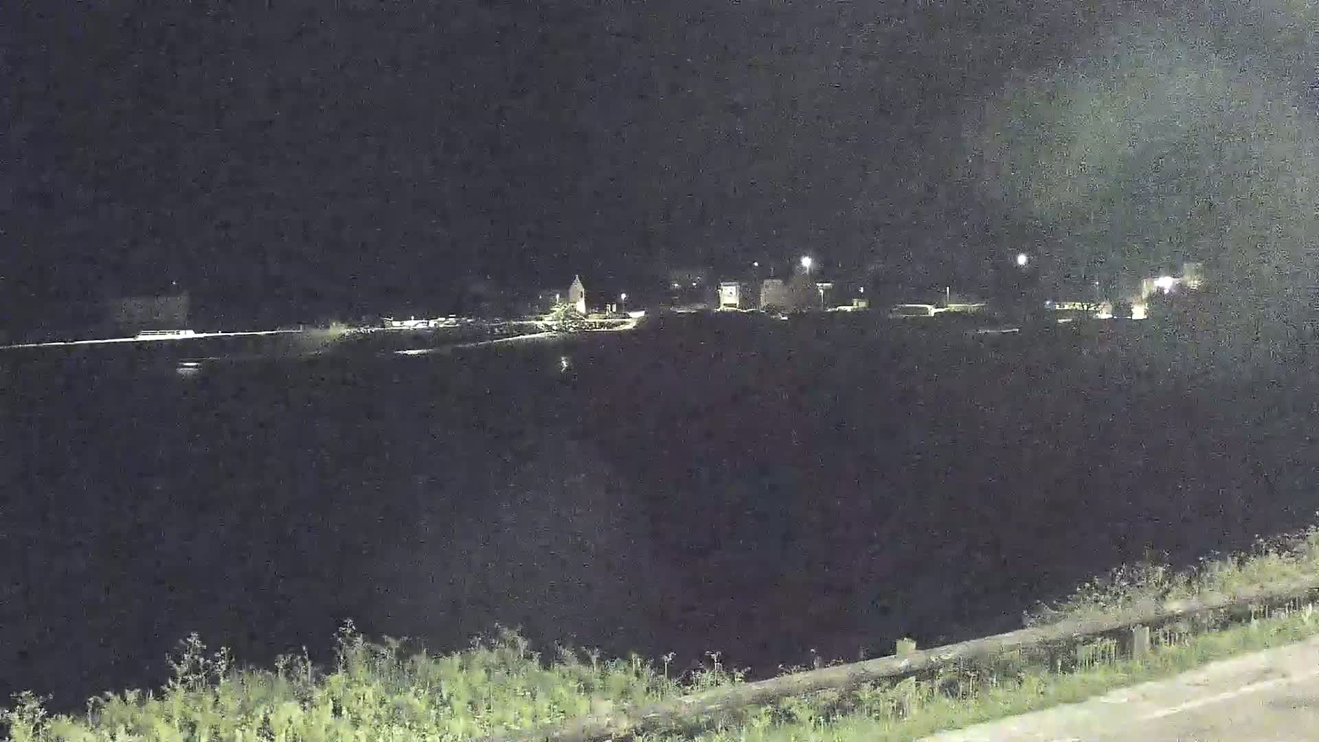 A nighttime view shows a dark body of water in the foreground with a dimly lit town visible in the distance under a dark sky.