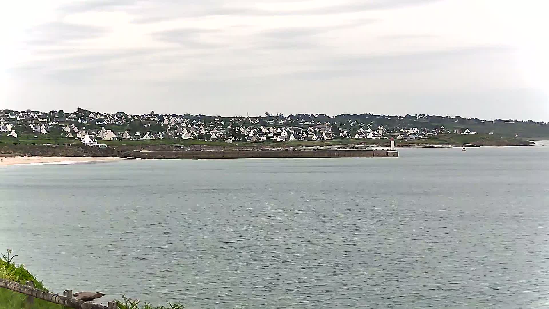 A coastal town with white houses sits on a hill overlooking a calm, gray sea under an overcast sky.
