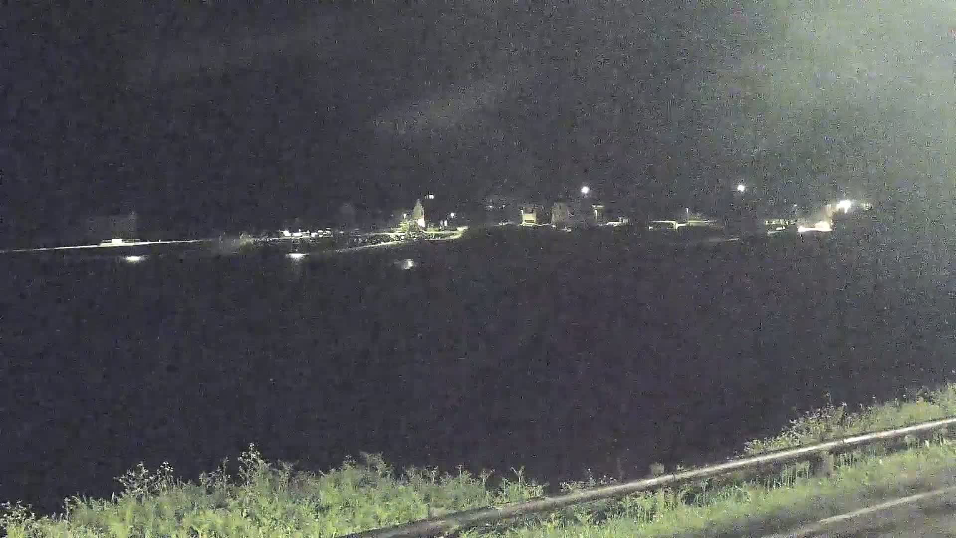 A nighttime view shows a dark body of water reflecting a few distant lights from a small town across the water, with vegetation visible in the foreground.