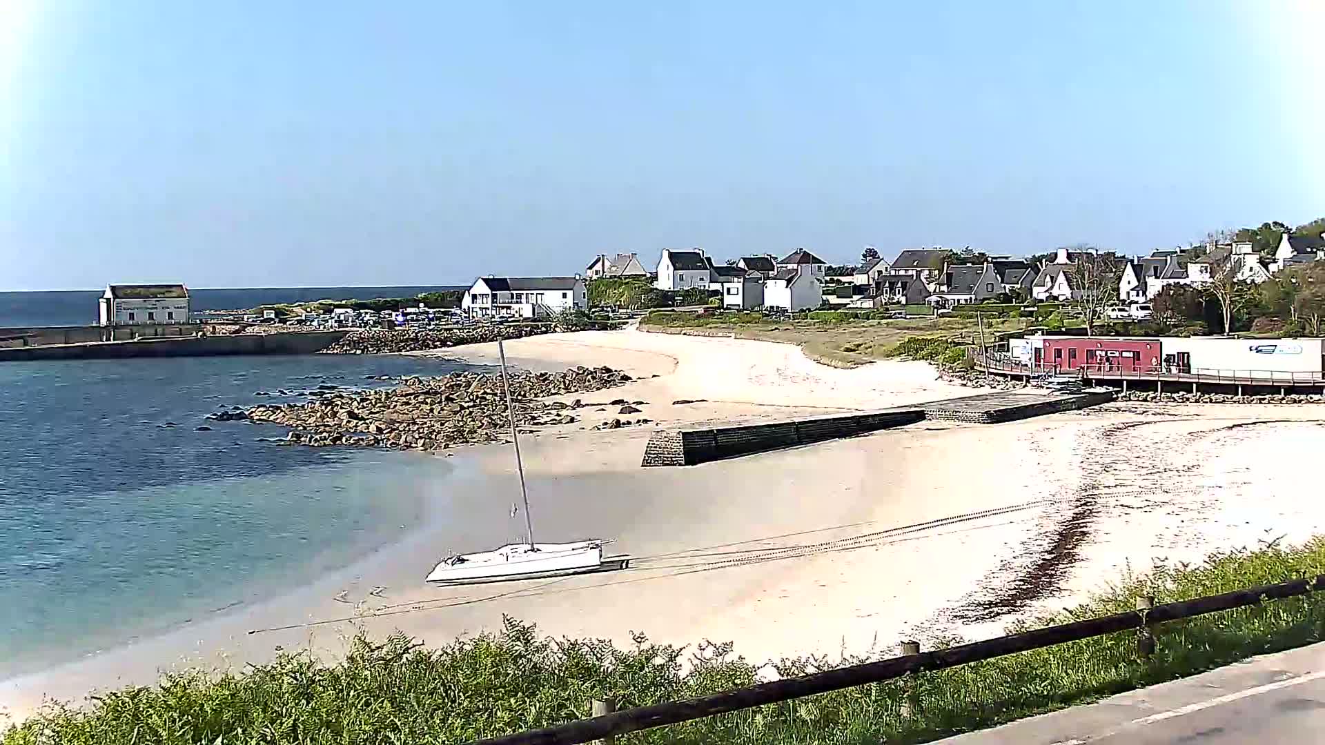 A small sailboat sits on a sandy beach next to calm, clear water, with a small town and buildings in the background under a clear blue sky.
