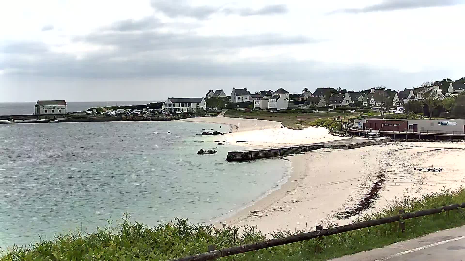 A small, sandy beach curves around a calm, light-green bay, with a cluster of houses and a small building on the shore under a cloudy sky.