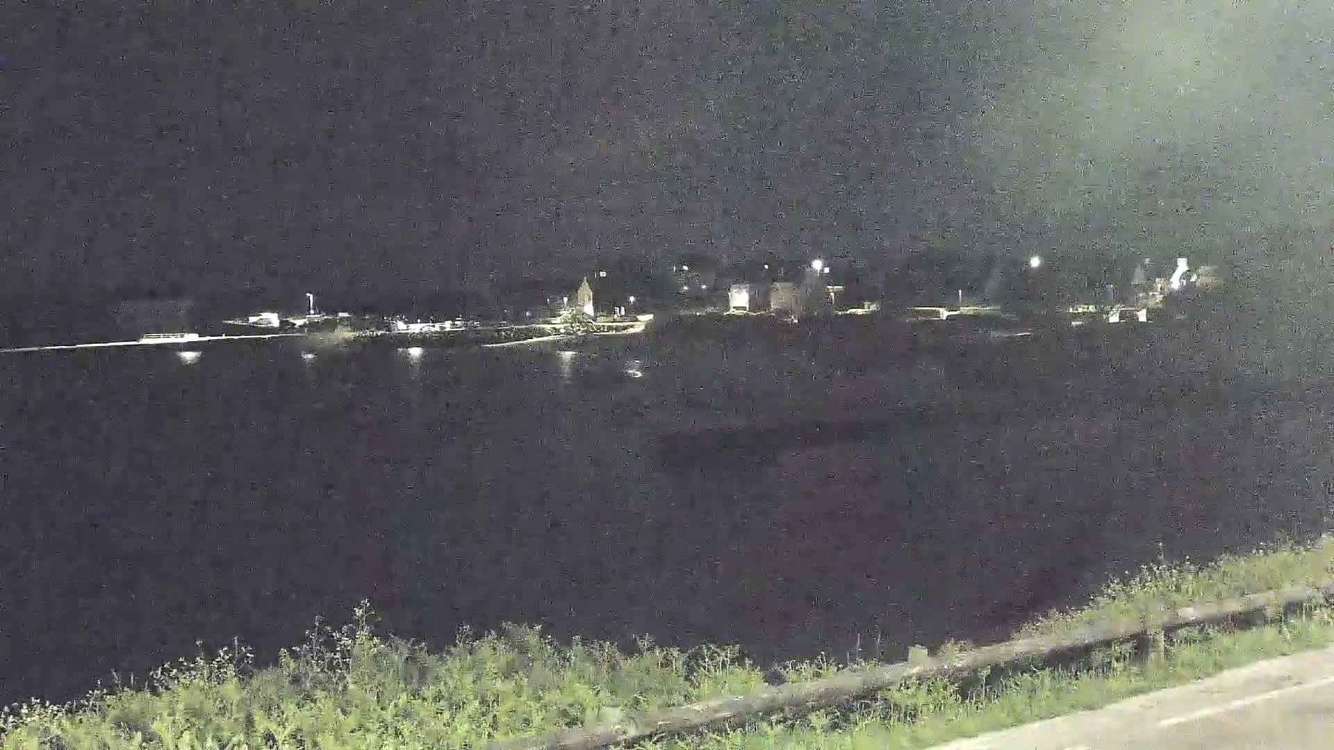 A nighttime view shows a body of dark water reflecting a few lights from a distant town across the water.