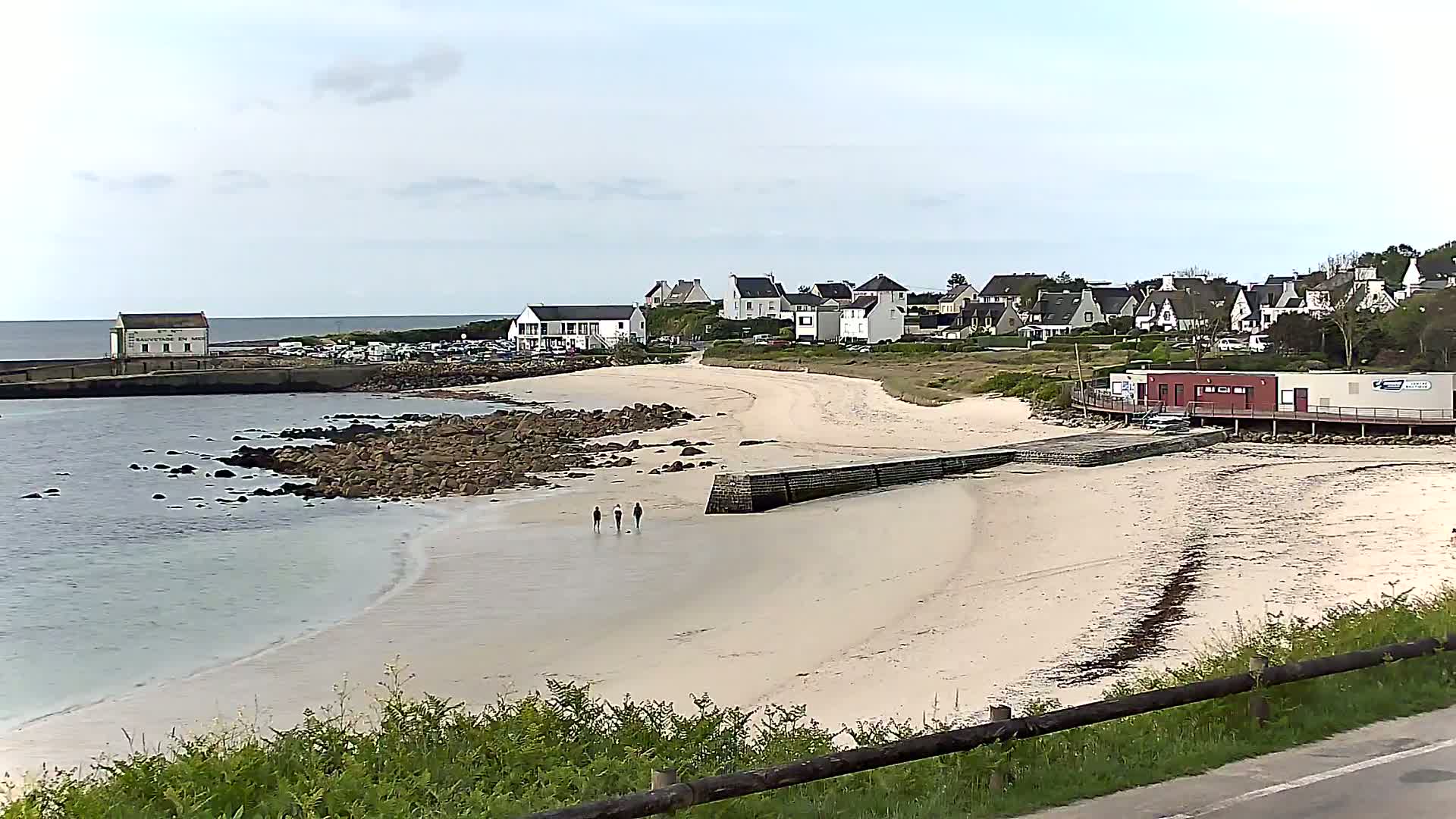 A sandy beach curves around a rocky shoreline, with a small village and a few people walking on the sand under a mostly cloudy sky.
