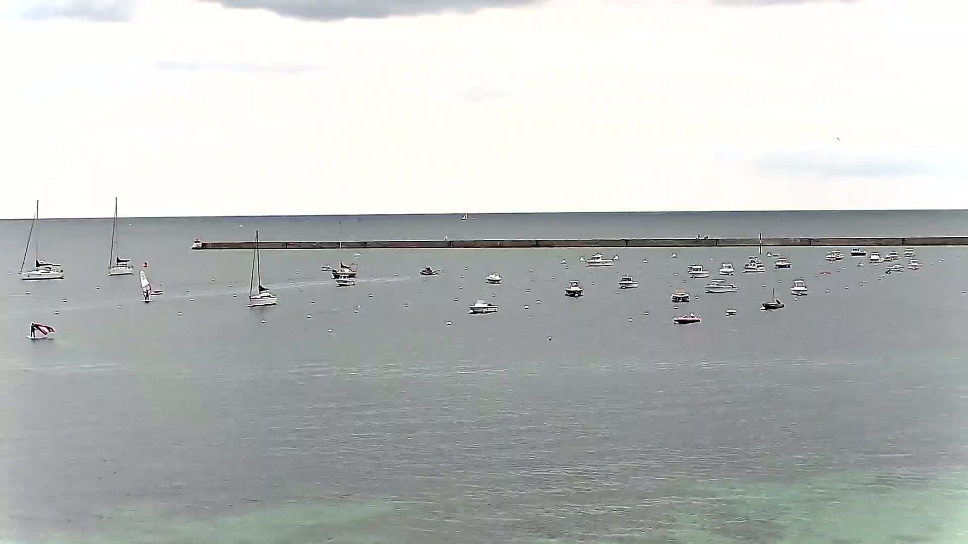 Numerous sailboats and motorboats are moored in calm, gray water behind a long breakwater under an overcast sky.