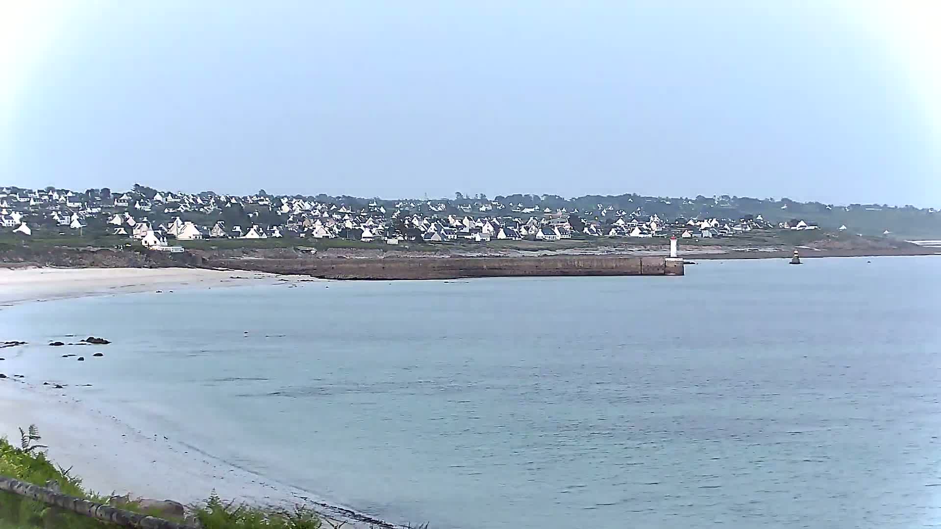 A coastal town with white houses sits atop a rocky shoreline overlooking a calm, light-blue ocean with a small lighthouse at a harbor entrance.
