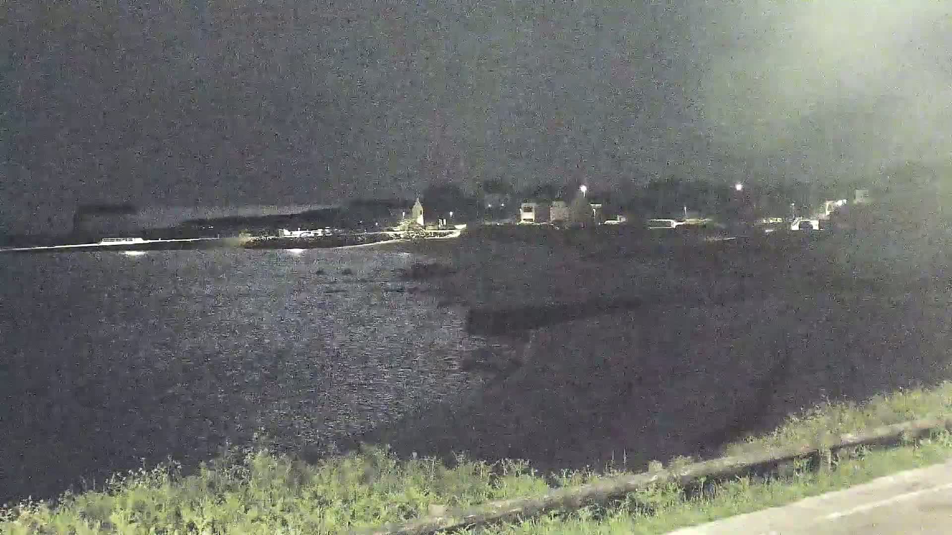 A nighttime view shows a dark body of water reflecting a few lights from a distant shoreline with some low-lying vegetation in the foreground.