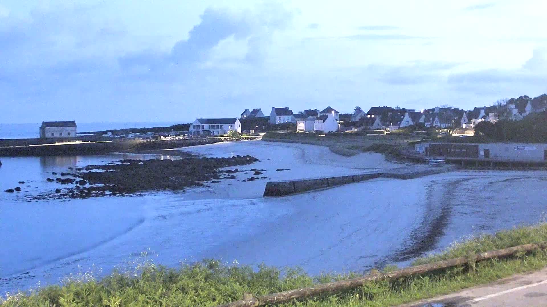 A small coastal village with white buildings sits on a sandy beach next to calm, shallow water under a mostly cloudy sky.