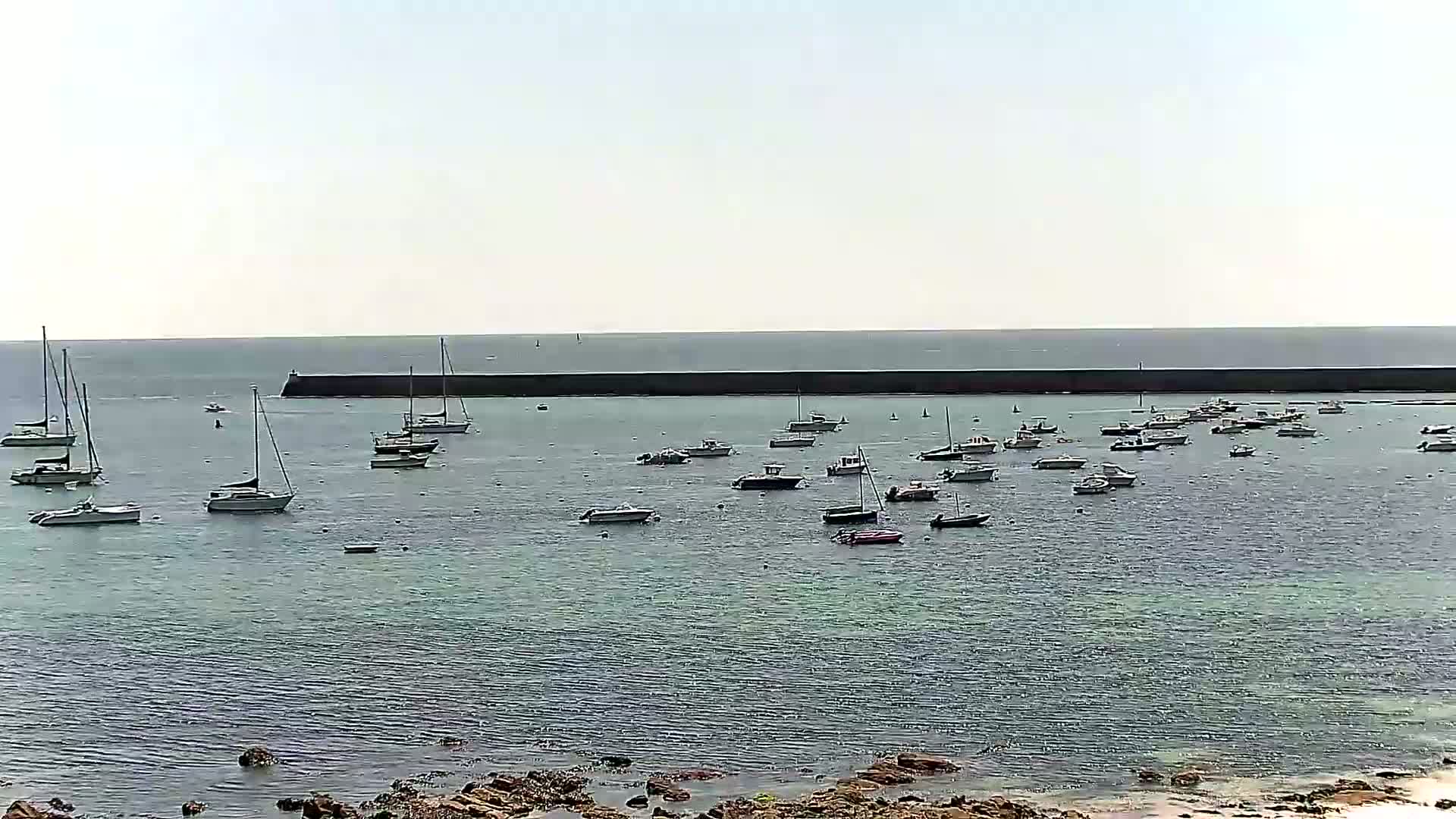 Numerous sailboats and motorboats are moored in calm water behind a long, dark breakwater on a bright, overcast day.