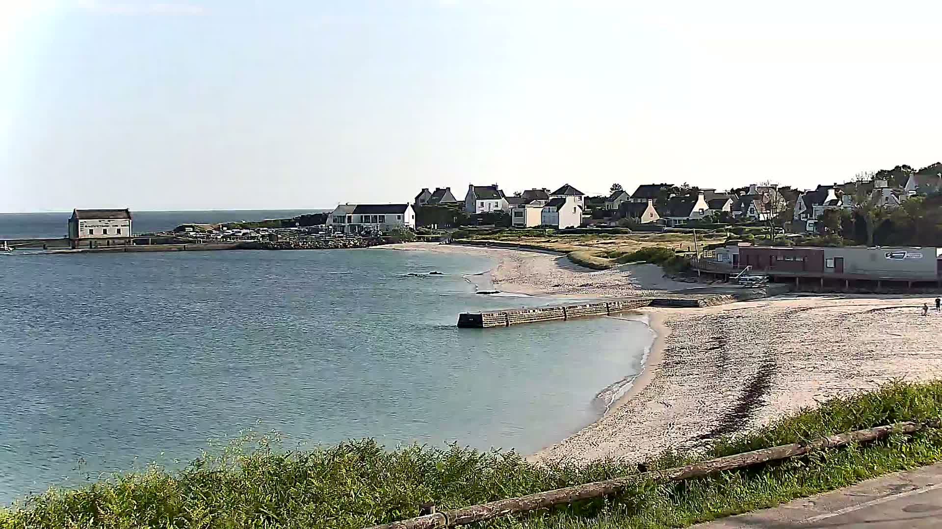 A sandy beach curves along a calm, light-blue body of water, with a small village of houses and buildings situated behind it on a sunny day.